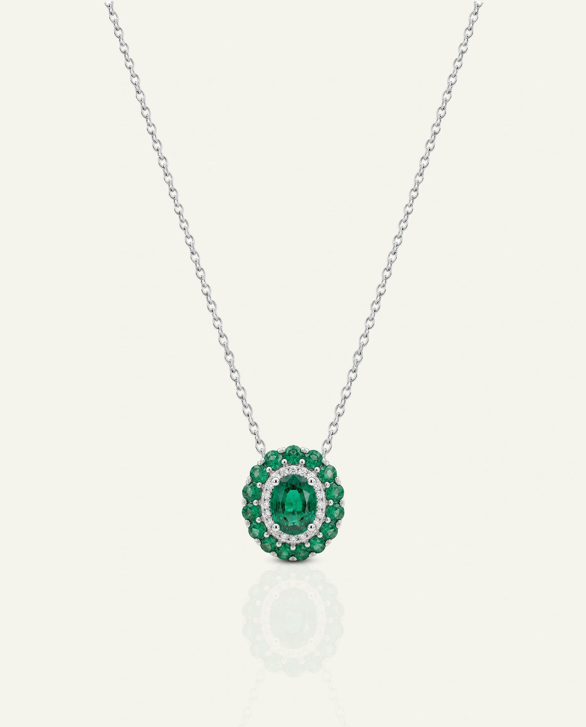 A silver chain necklace with a circular pendant featuring a central oval green emerald surrounded by a halo of round green stones and small clear stones, shown on a light cream background.