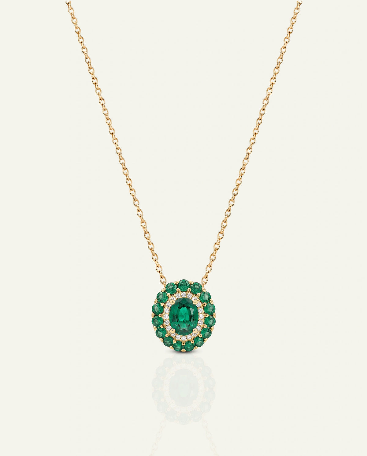 A gold chain necklace with a circular pendant featuring a central oval green emerald and a surrounding halo of round green and clear stones, shown on a light cream background.