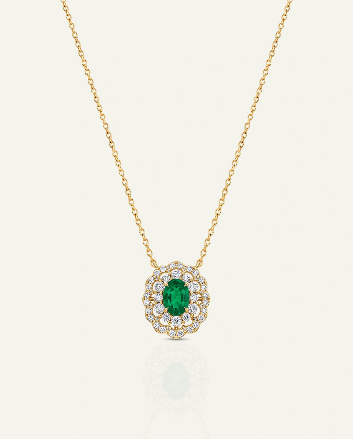 A gold chain necklace featuring a circular pendant with a central green emerald-cut stone surrounded by a halo of small clear stones, shown on a light cream background.