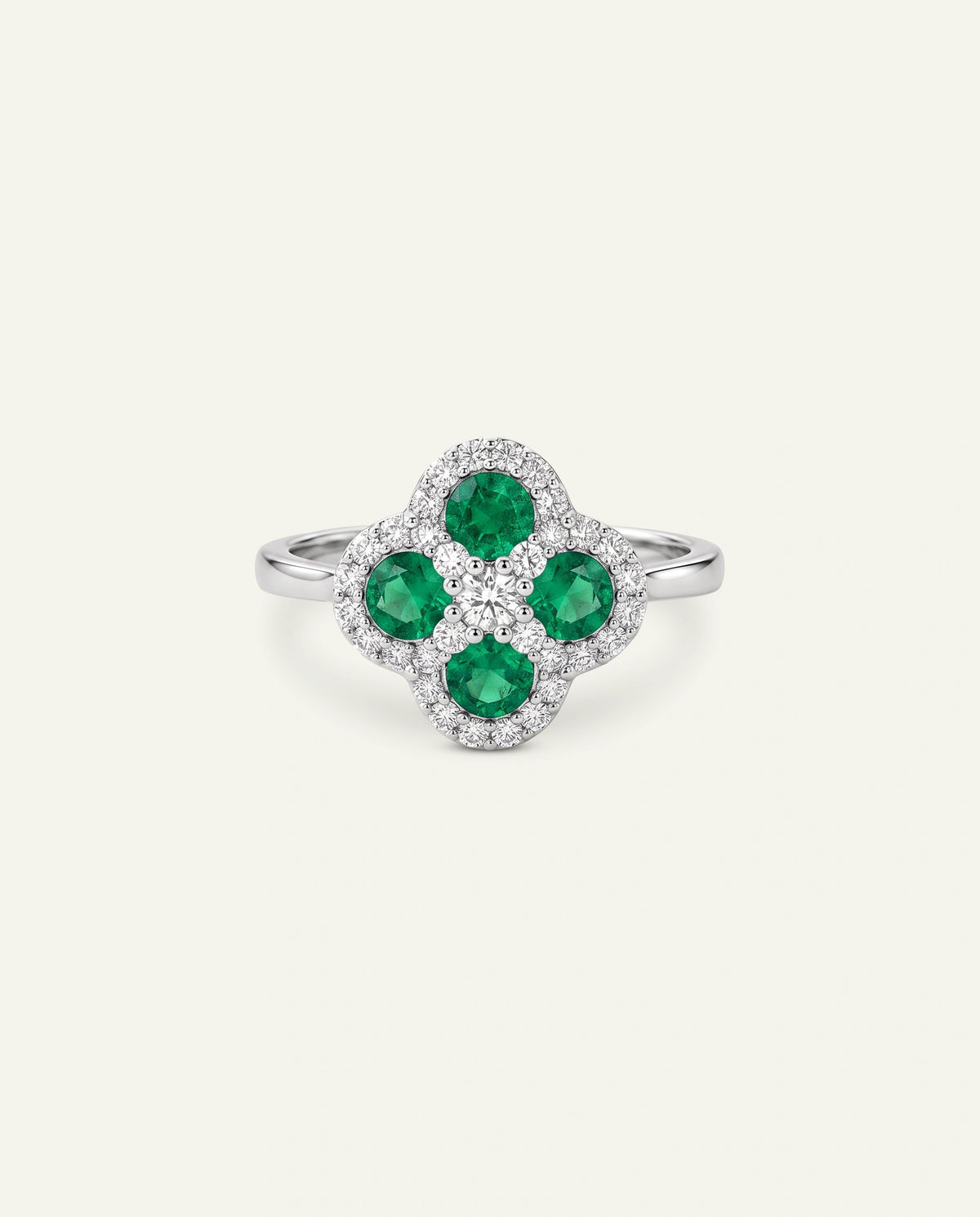 A silver four-petal flower ring featuring three round green emerald-cut center stones and a halo of small clear pavÃ©-set stones, shown on a light cream background.