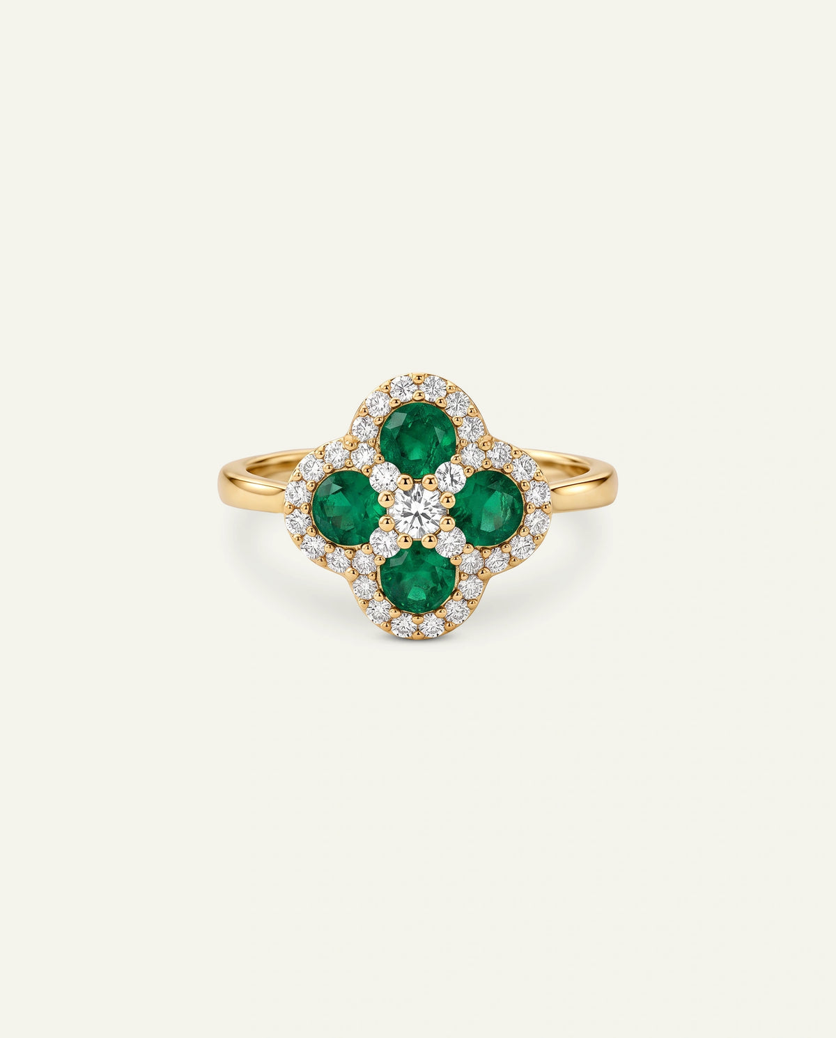 A yellow gold ring featuring a four-petal clover motif with round green emeralds and a halo of small clear pavÃ© stones, set against a solid off-white background.