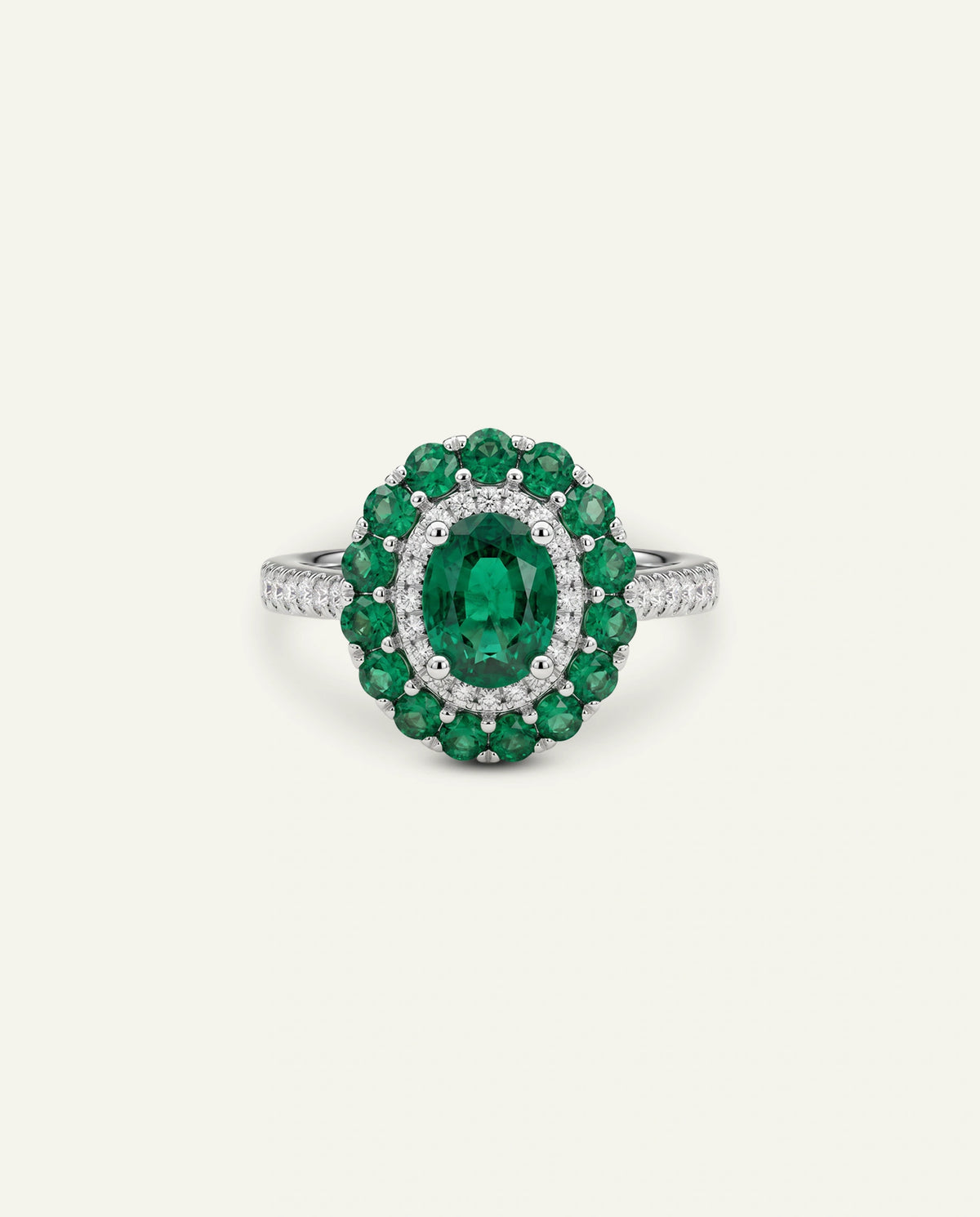 An oval emerald-cut green gemstone ring with a halo of round green stones and small clear accent stones on a white metal band, centered on a light cream background.