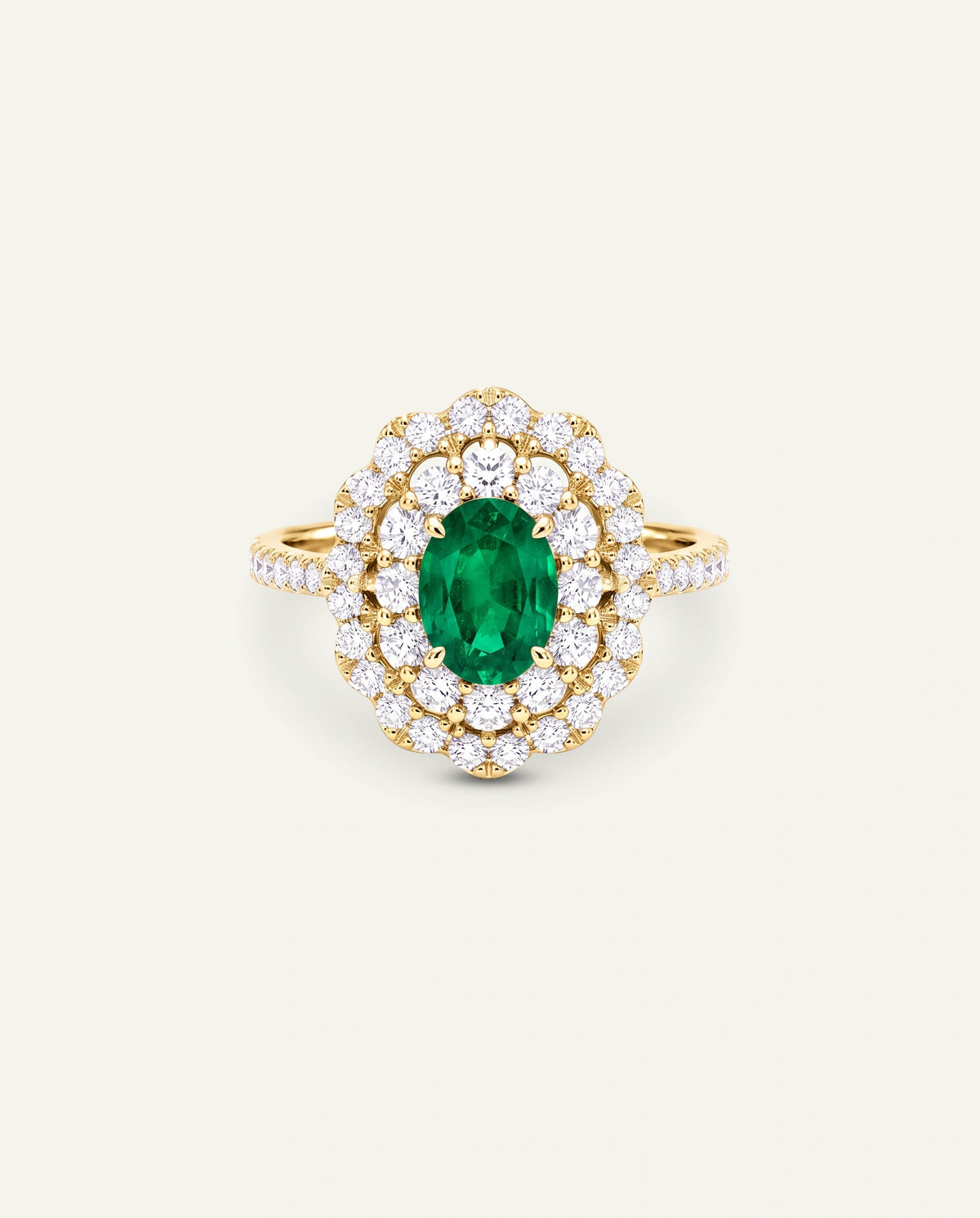 A yellow gold ring featuring a central oval-cut green emerald gemstone surrounded by a double halo of round brilliant-cut diamonds, with the band decorated with small side diamonds, set against a solid off-white background.