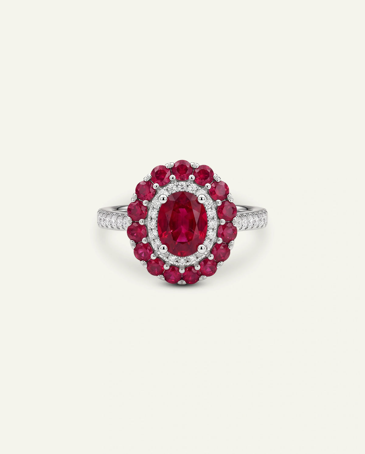 A silver-toned ring featuring a large oval red ruby center stone surrounded by a double halo of round red rubies and small clear stones, with a diamond-like pavÃ© band, set against a light cream background.