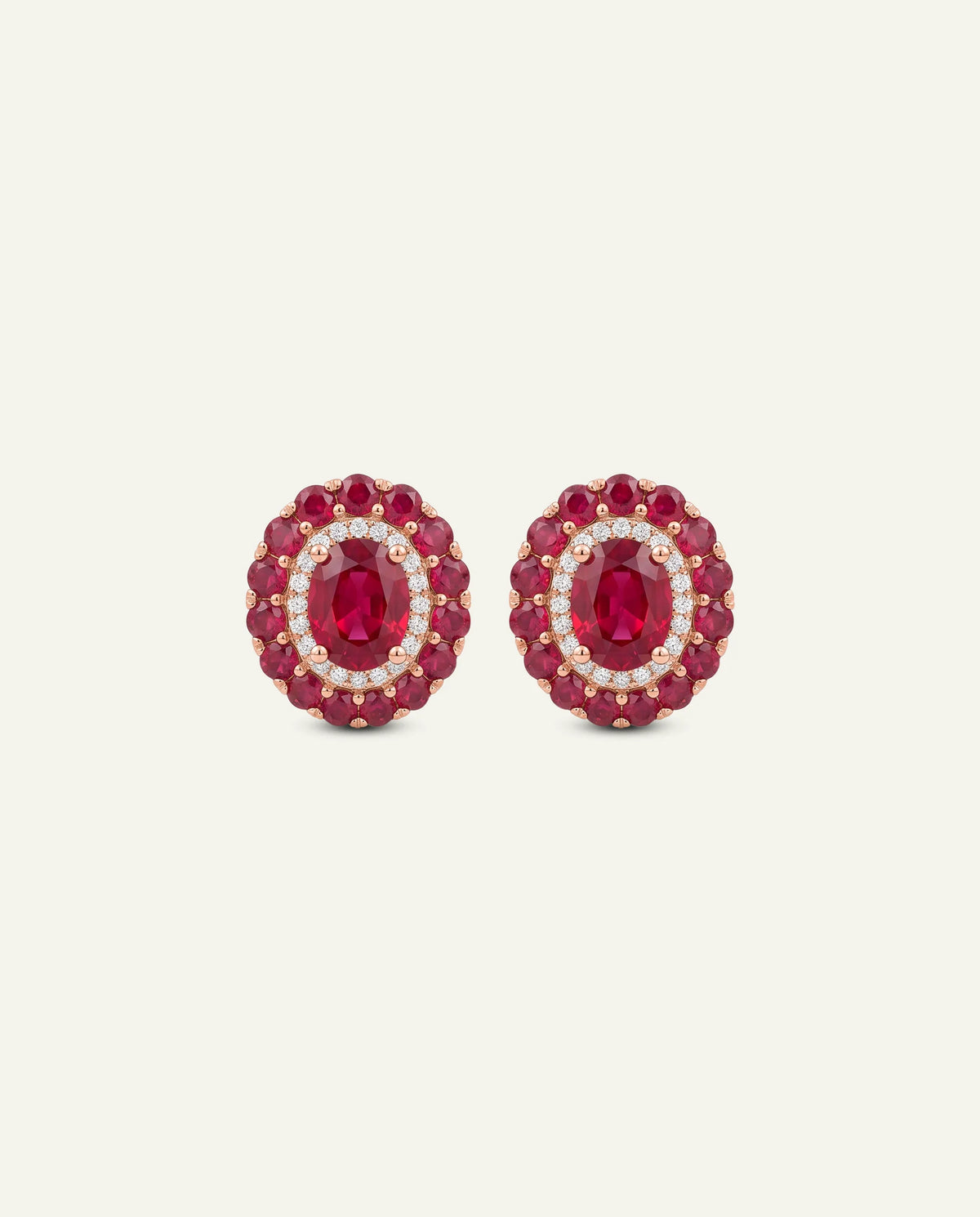A pair of oval stud earrings featuring a central oval-cut deep red ruby surrounded by a halo of small clear stones, with a border of larger round red stones, set in yellow gold on a light cream background.
