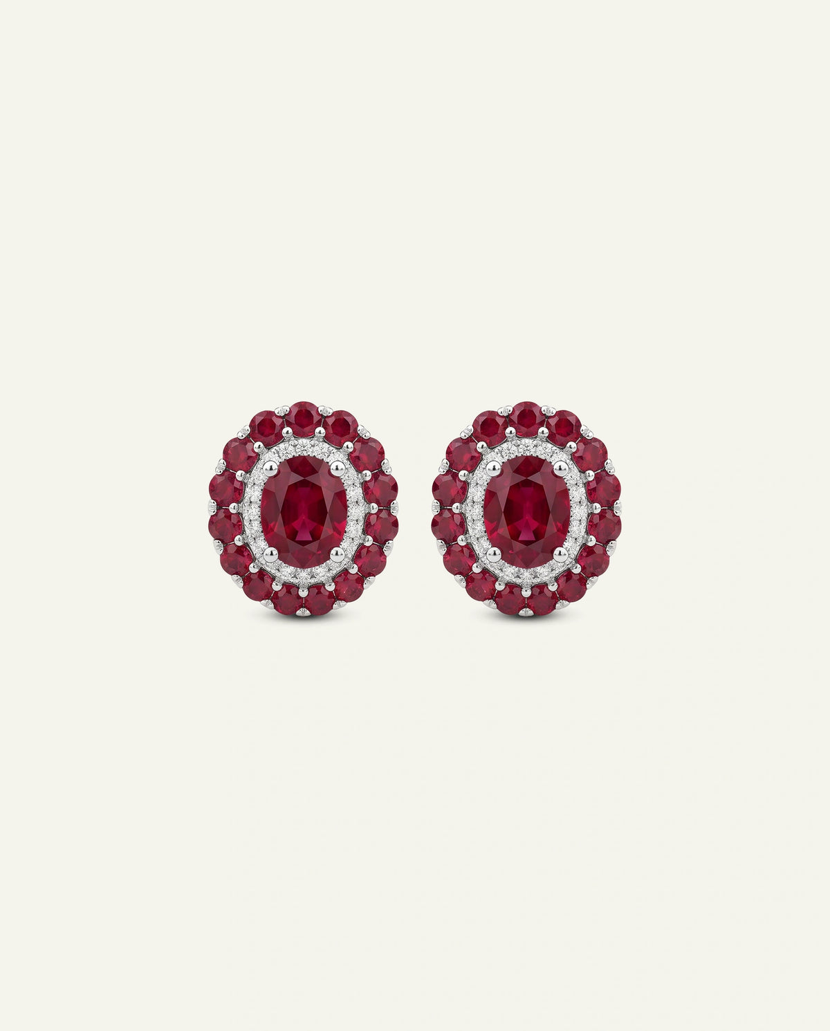 A pair of oval stud earrings featuring oval-cut deep red ruby centers surrounded by a halo of small clear stones, with a border of round red stones, set against a light cream background.