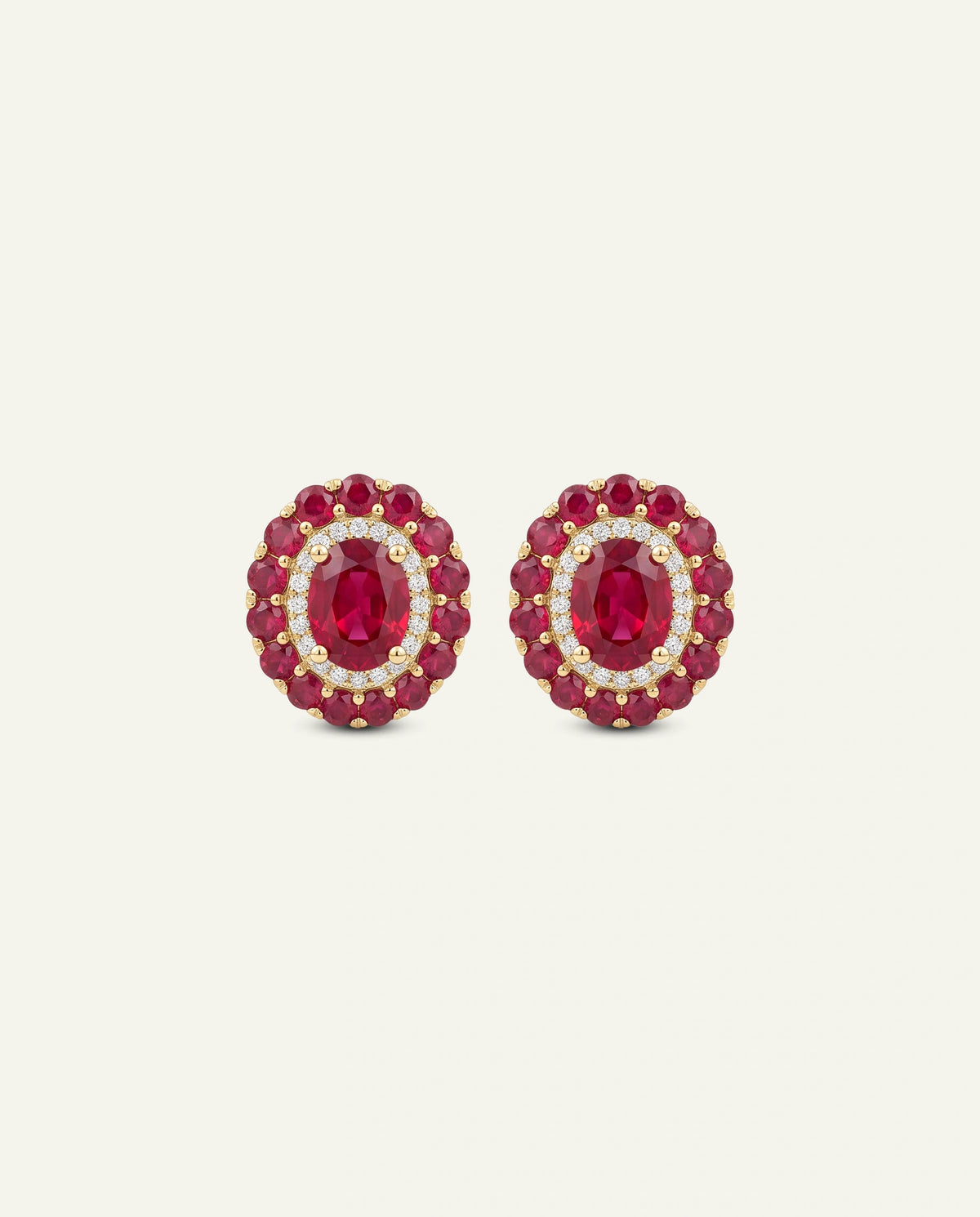 A pair of oval stud earrings featuring deep red oval-cut center stones surrounded by a halo of small clear stones and a border of red stones, set in yellow gold, on a light cream background.