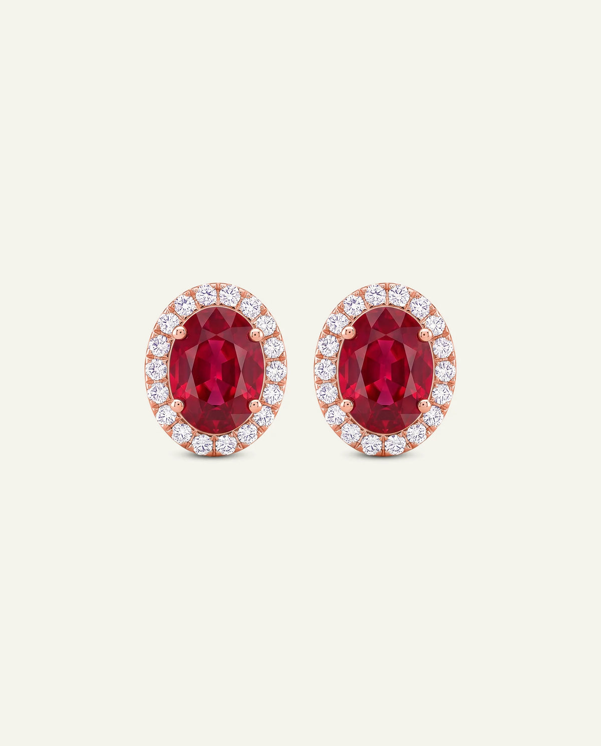 A pair of oval stud earrings featuring a deep red center stone surrounded by a halo of small clear stones in rose gold settings, presented on a light cream background.
