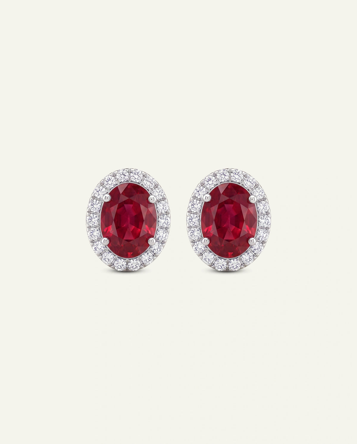 A pair of oval stud earrings featuring deep red oval-cut center stones surrounded by a halo of small clear stones in a silver-toned setting, shown on a plain light beige background.