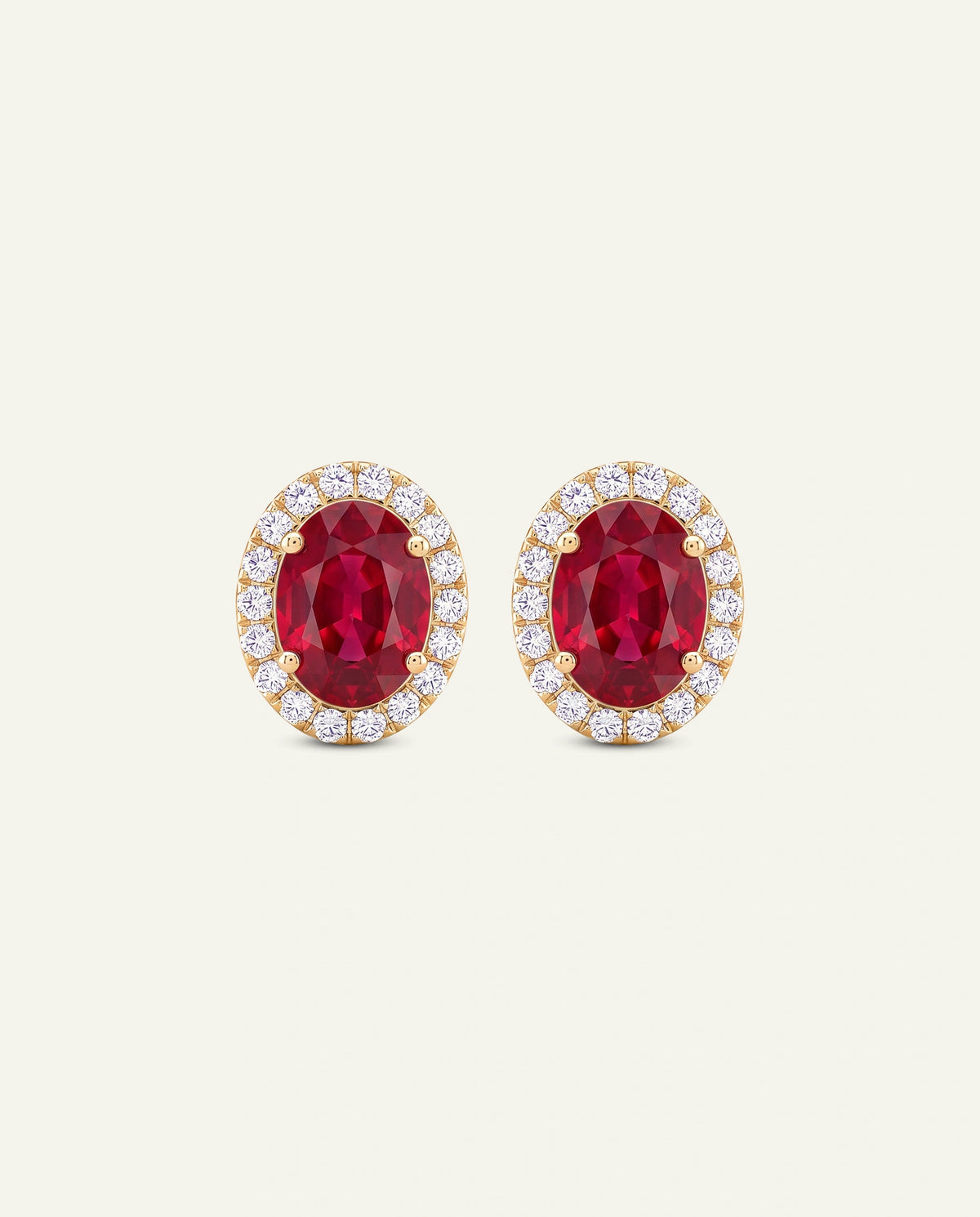 A pair of oval stud earrings featuring a deep red central stone surrounded by a halo of small clear stones in a yellow gold setting, shown on a light cream background.