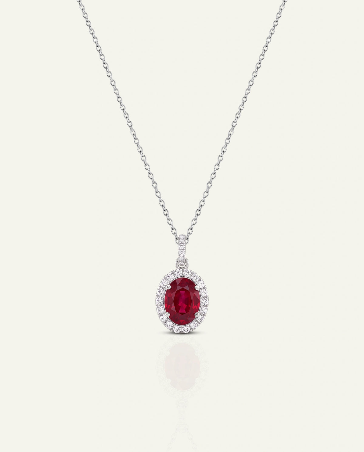 An elegant oval pendant necklace featuring a vibrant red ruby center stone surrounded by a halo of small clear diamonds, suspended on a delicate silver-toned chain against a light cream background.