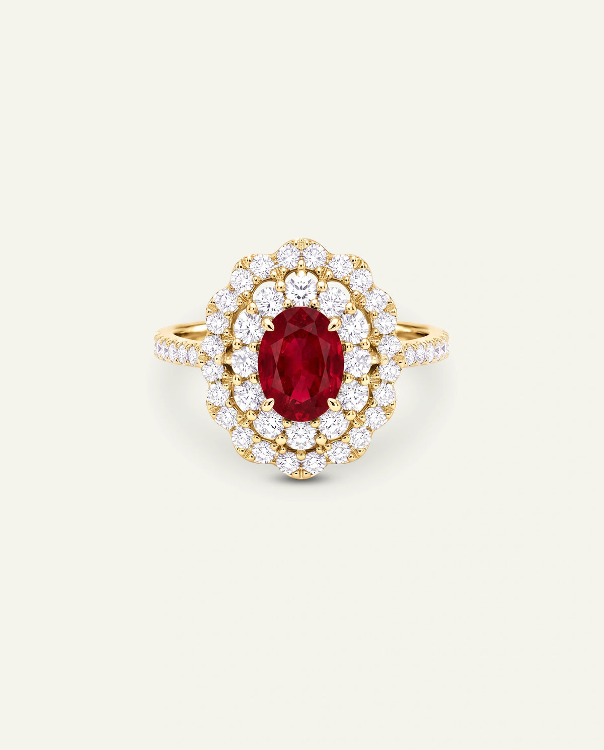 A yellow gold ring featuring an oval-cut red ruby center stone surrounded by a double halo of round brilliant-cut diamonds, with the band adorned with small pavÃ© diamonds, set against a light off-white background.