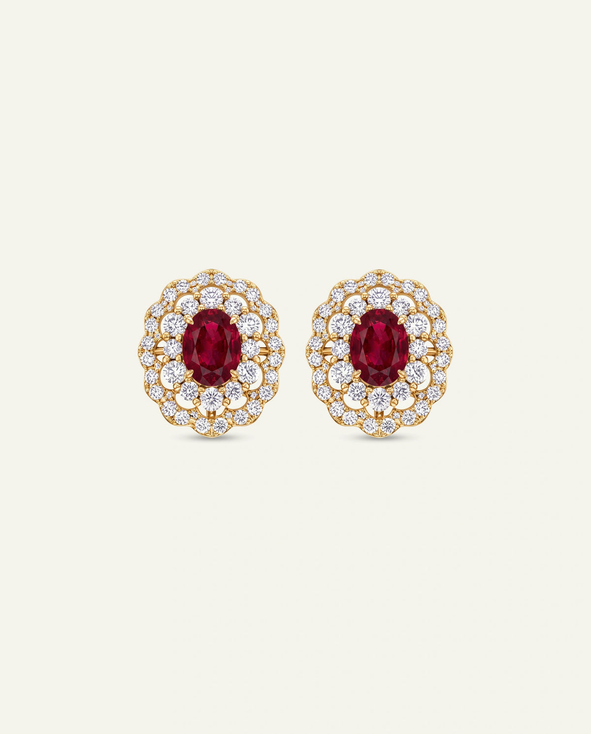 A pair of yellow gold stud earrings featuring central oval-cut red rubies framed by a intricate, two-tier scalloped halo of small round clear pavé diamonds, set against an off-white background.