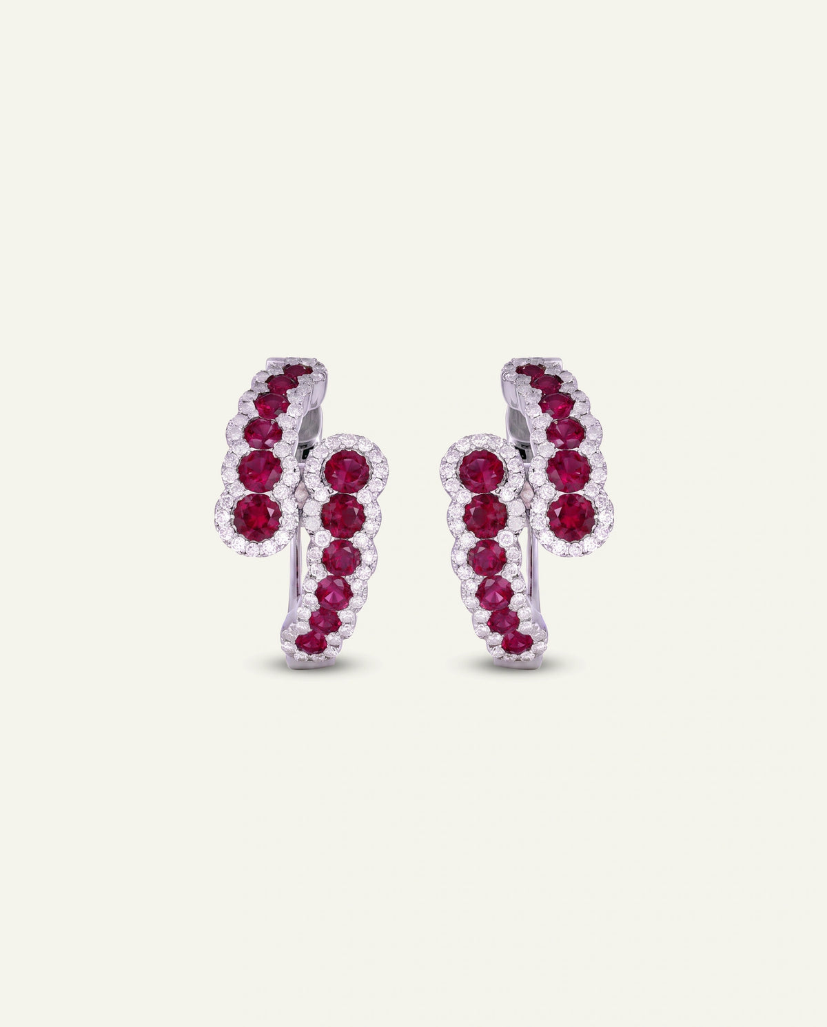 A pair of silver-toned huggie hoop earrings embellished with rows of round-cut deep red rubies and small clear stones, set against a solid off-white background.