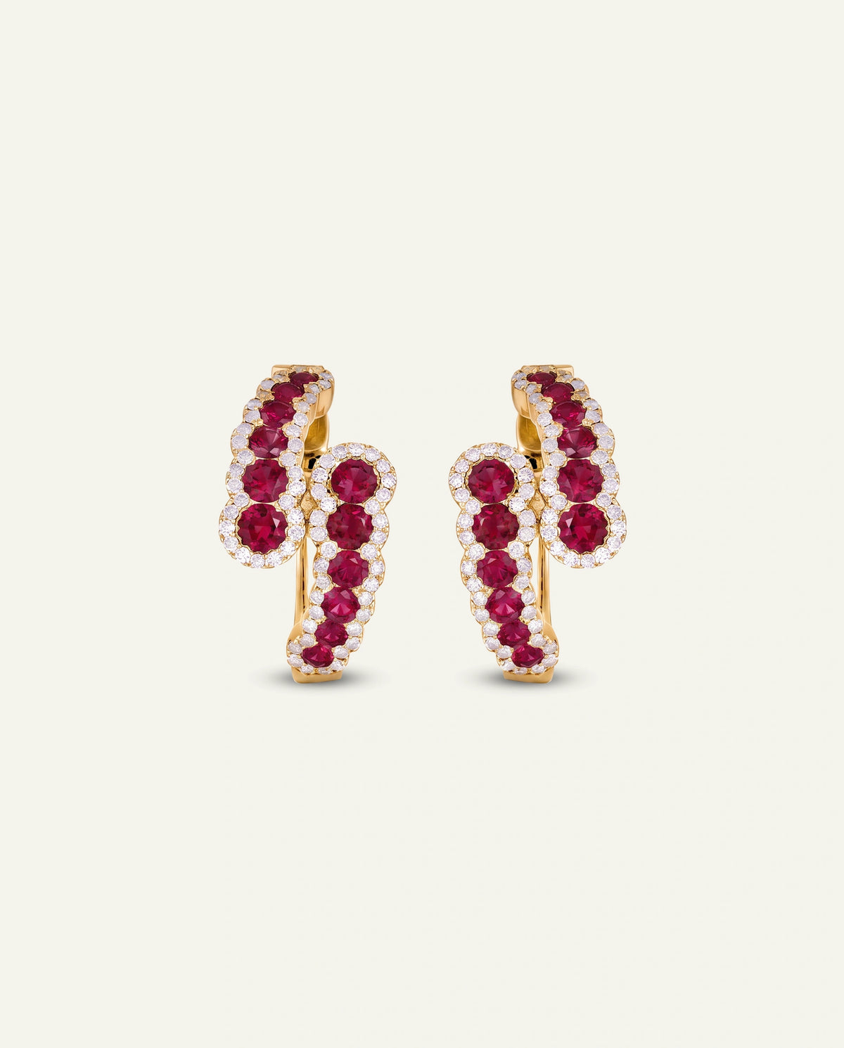 A pair of gold huggie hoop earrings adorned with rows of round-cut deep red rubies and small clear stones, set against a solid off-white background.