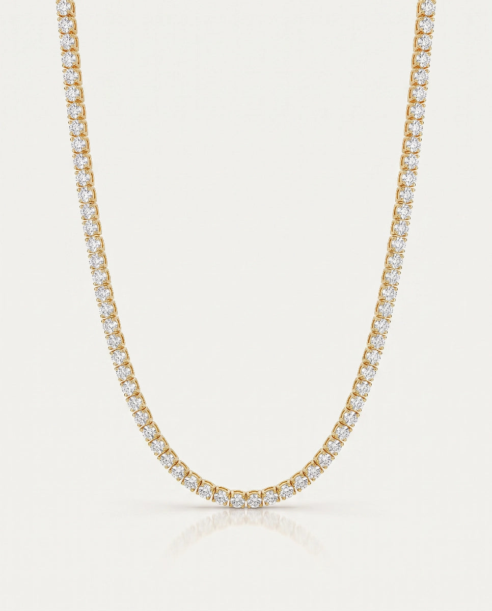A yellow gold tennis necklace paved with small clear stones, shown against a light cream background with a soft reflection below.