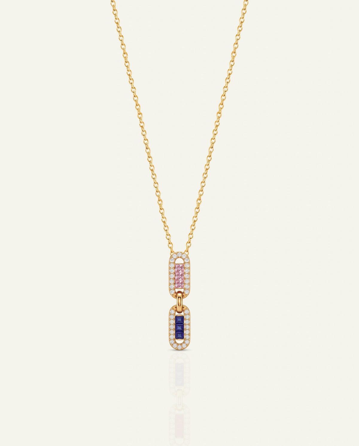 A delicate gold chain necklace featuring a double-drop pendant. The top drop is paved with small pink gemstones, and the bottom drop is paved with small clear stones surrounding a central rectangular blue gemstone, all set in yellow gold against a light cream background.