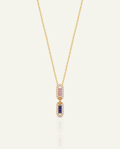 A delicate gold chain necklace featuring a double-drop pendant. The top drop is paved with small pink gemstones, and the bottom drop is paved with small clear stones surrounding a central rectangular blue gemstone, all set in yellow gold against a light cream background.