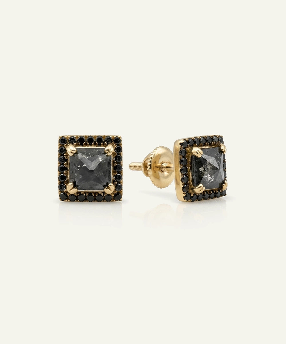 A pair of gold square stud earrings featuring a central dark gray gemstone surrounded by a halo of small black stones, shown on a light cream background.