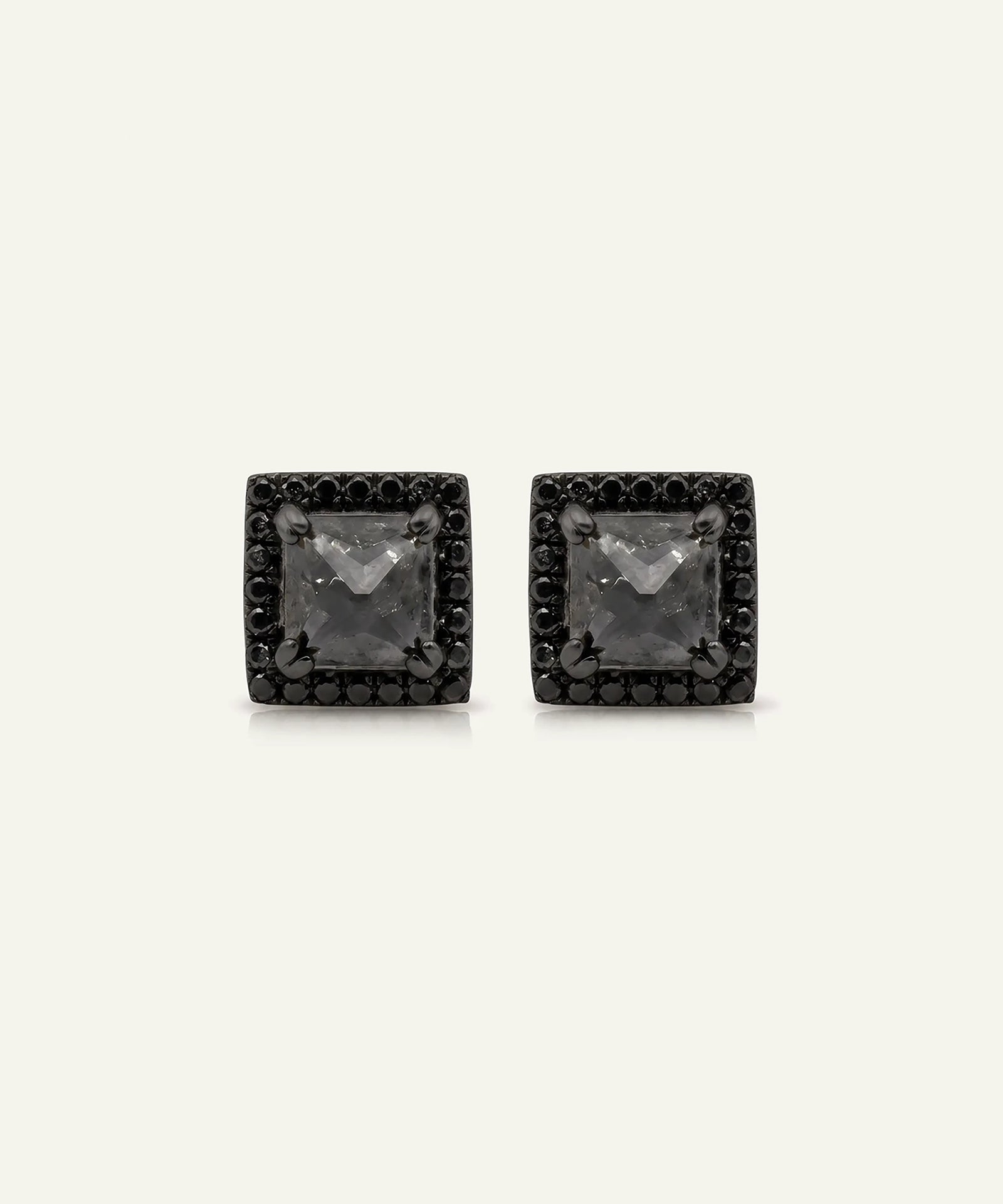 A pair of square stud earrings featuring a central clear gemstone surrounded by a halo of small black stones, set in a dark metal, shown on a light cream background.