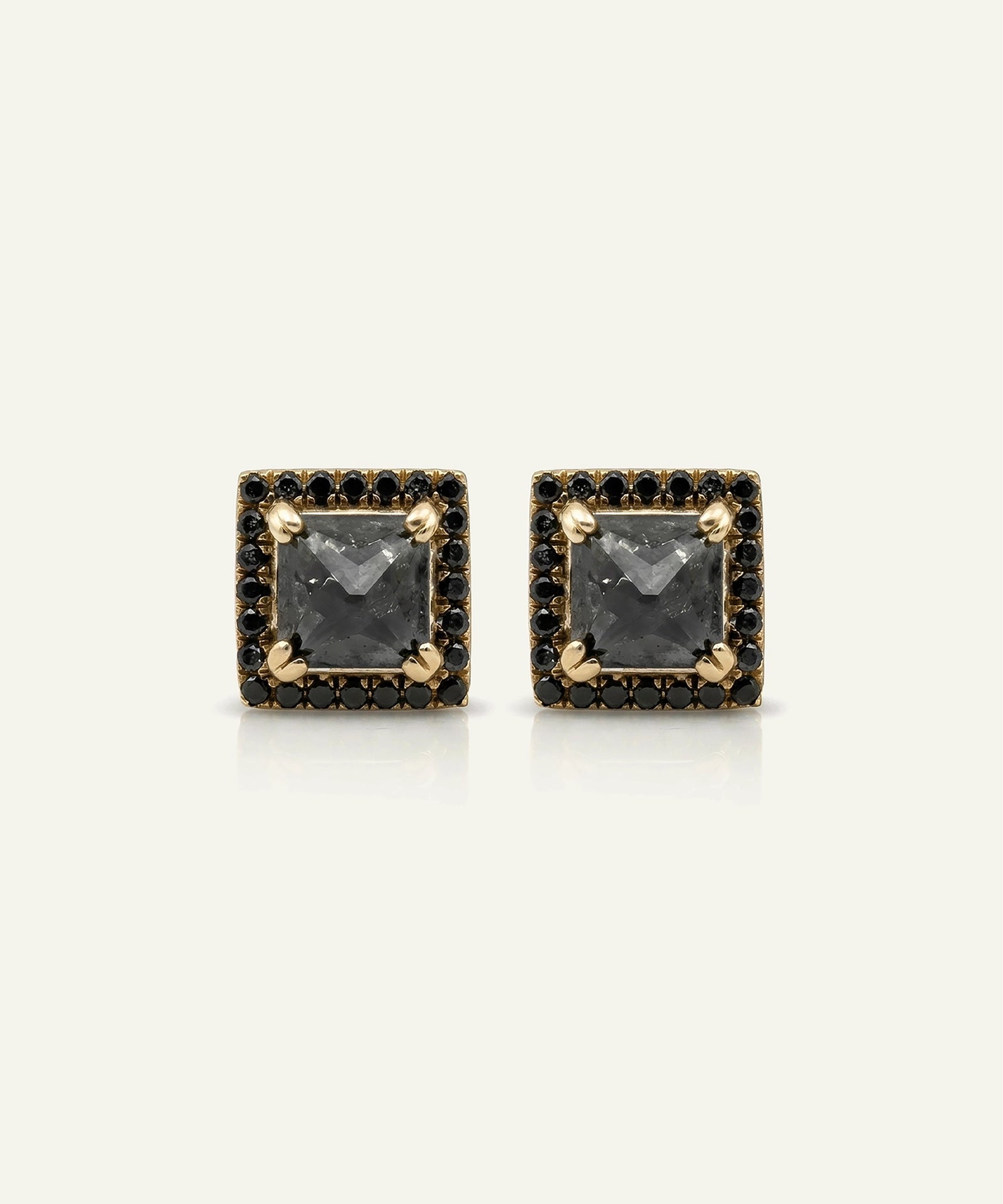 A pair of square stud earrings featuring a central dark gray stone surrounded by a halo of small black stones and accented with gold at the corners, set against a light off-white background.
