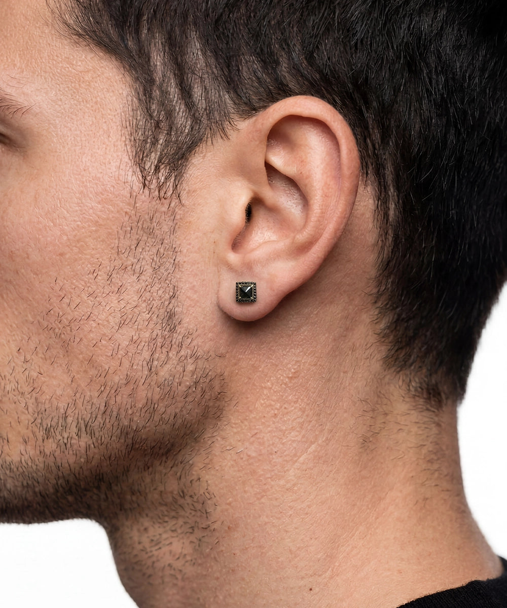 A close-up side profile of a man's ear wearing a small round stud earring with a dark green center and a gold halo setting, against a plain white background.