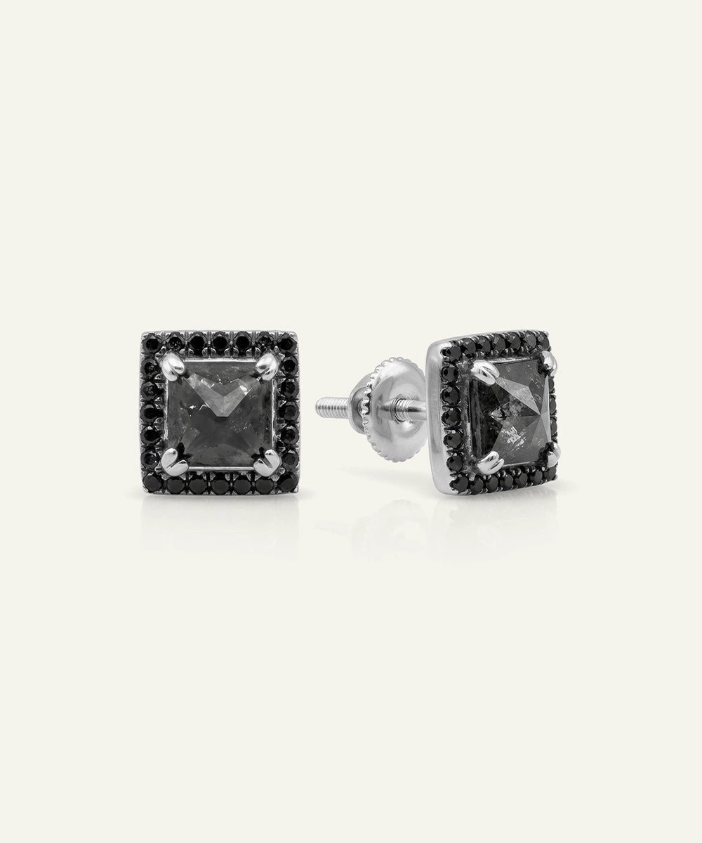 A pair of square stud earrings featuring a central dark gray gemstone surrounded by a halo of small black stones, set in silver-tone metal, shown on a light cream background.