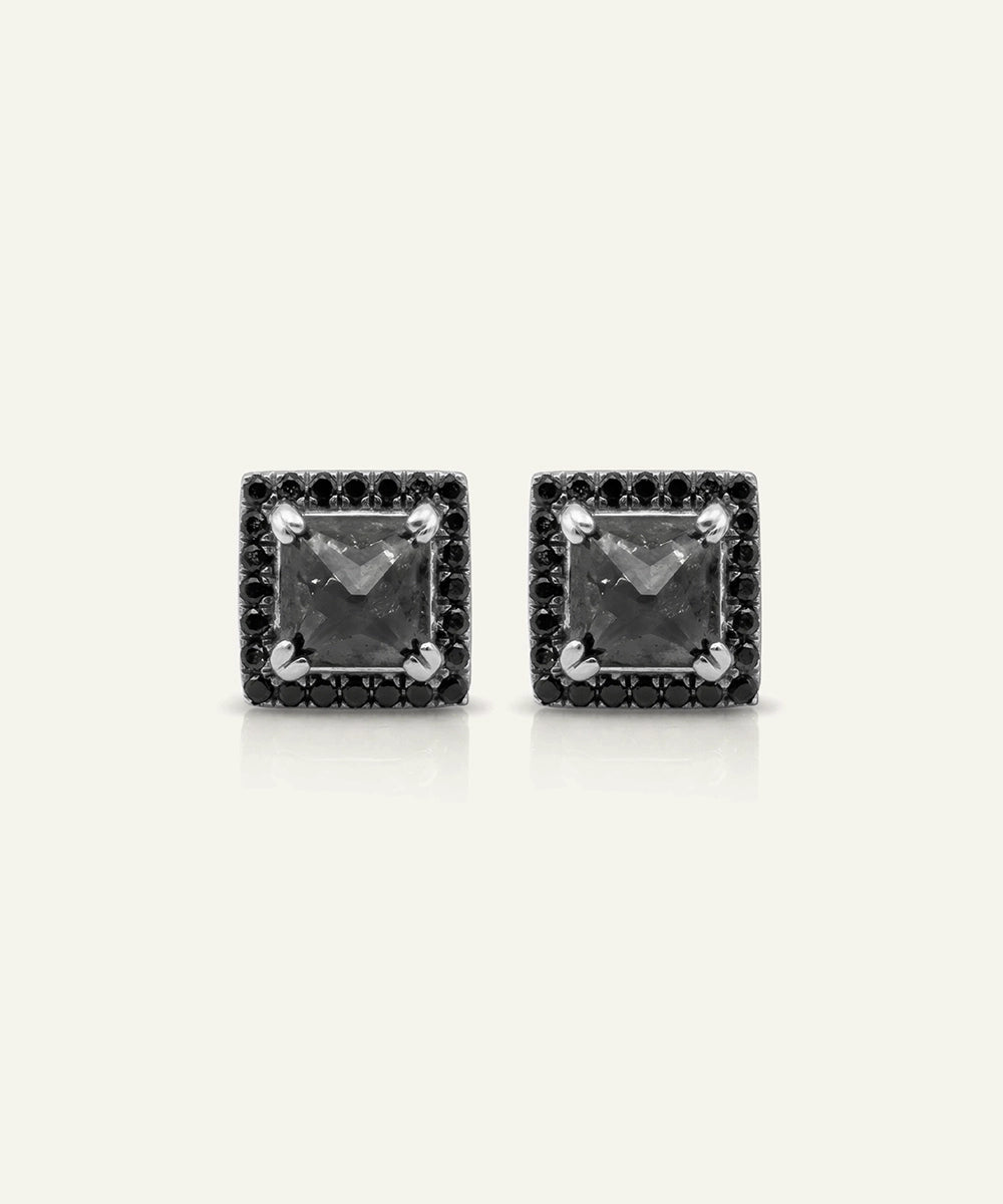 A pair of square stud earrings featuring a central square gray stone surrounded by a halo of small black stones, set in a silver-toned metal, on a light cream background.