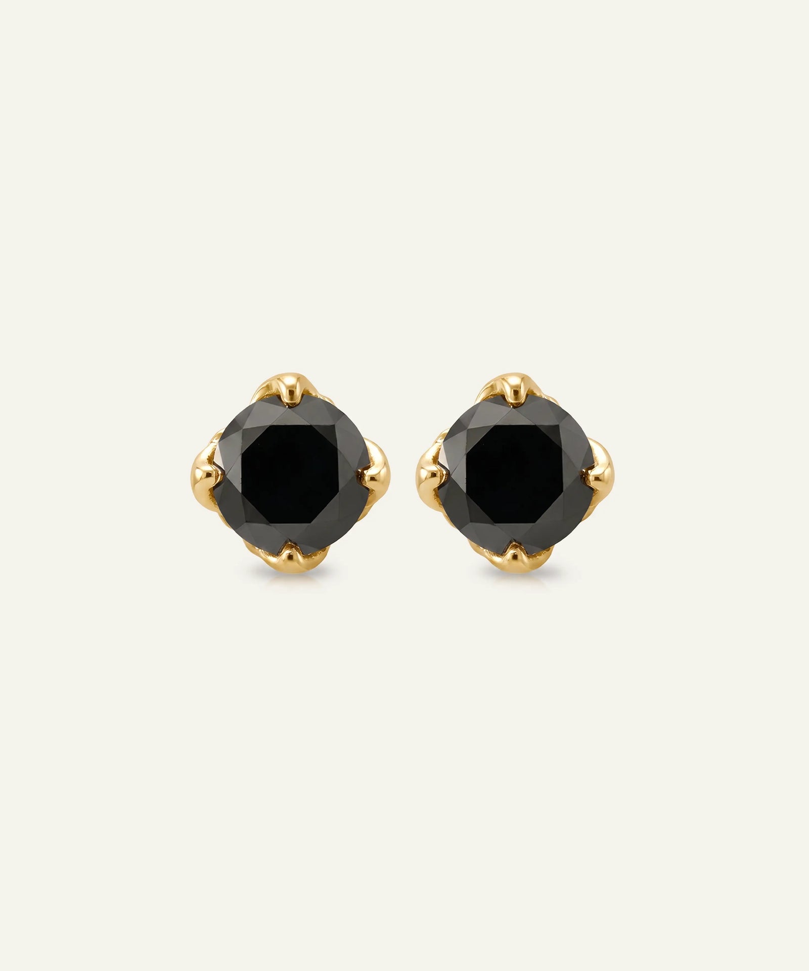 A pair of gold stud earrings with faceted black gemstones in a four-prong setting, shown on a light off-white background.