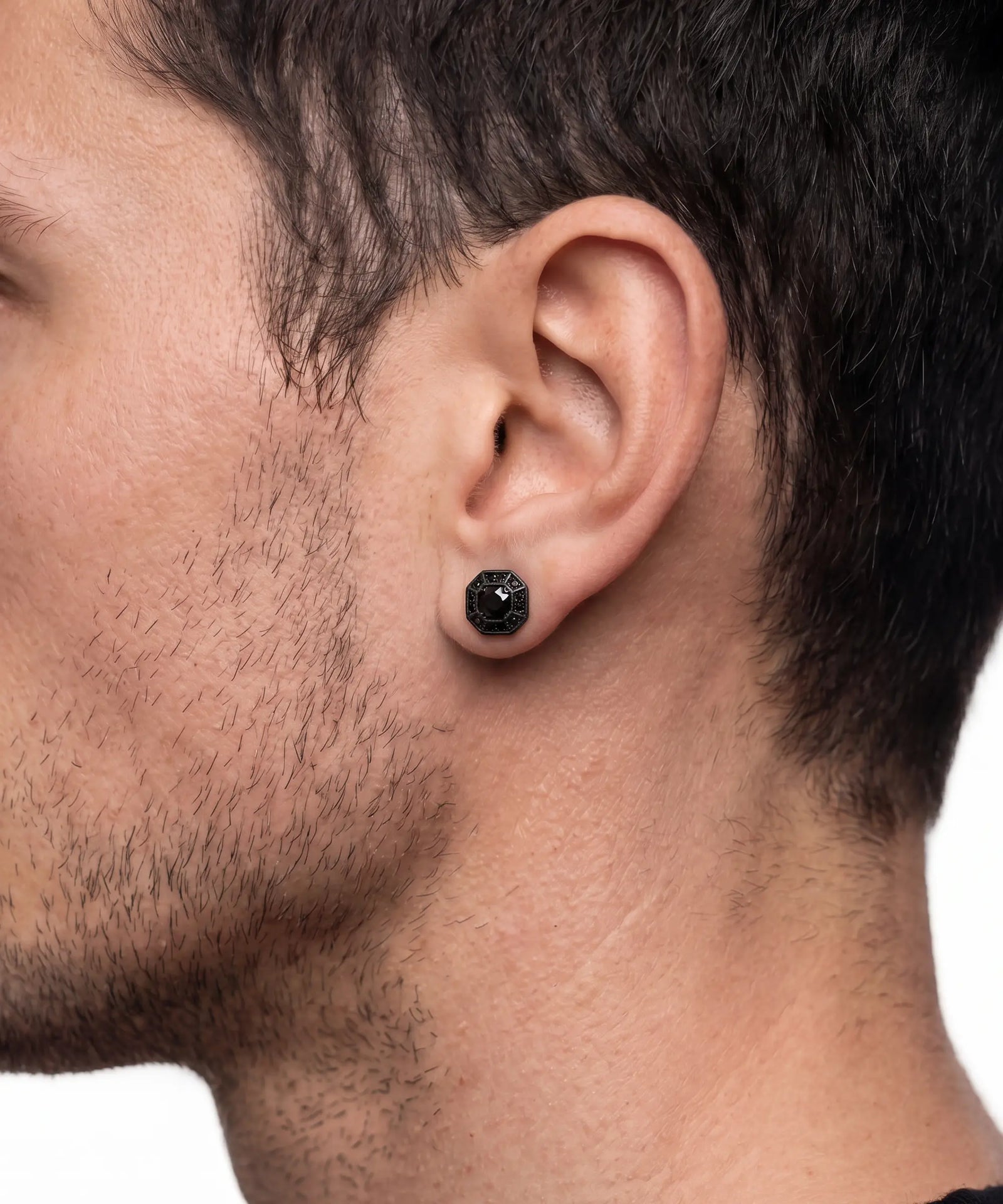 A close-up side profile of a man's head showing a black textured stud earring in his earlobe.