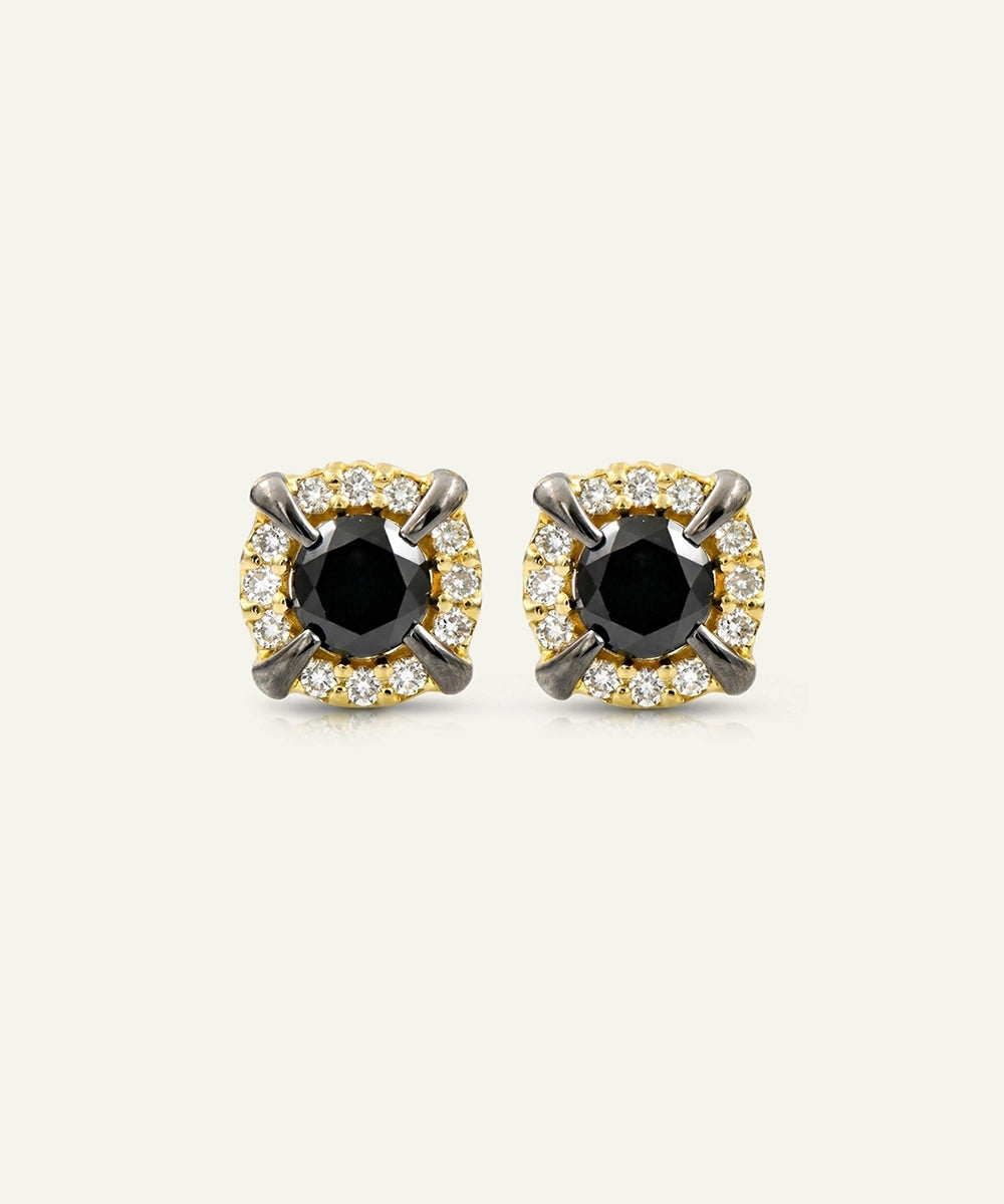 A pair of round stud earrings featuring a central black stone surrounded by a halo of small clear stones and four prongs with dark metallic accents, set against a plain off-white background.