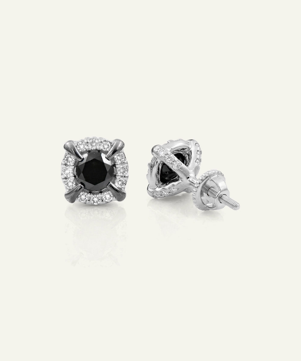 A pair of silver-toned stud earrings with a central round black gemstone surrounded by a halo of small clear stones, shown on a light cream background.