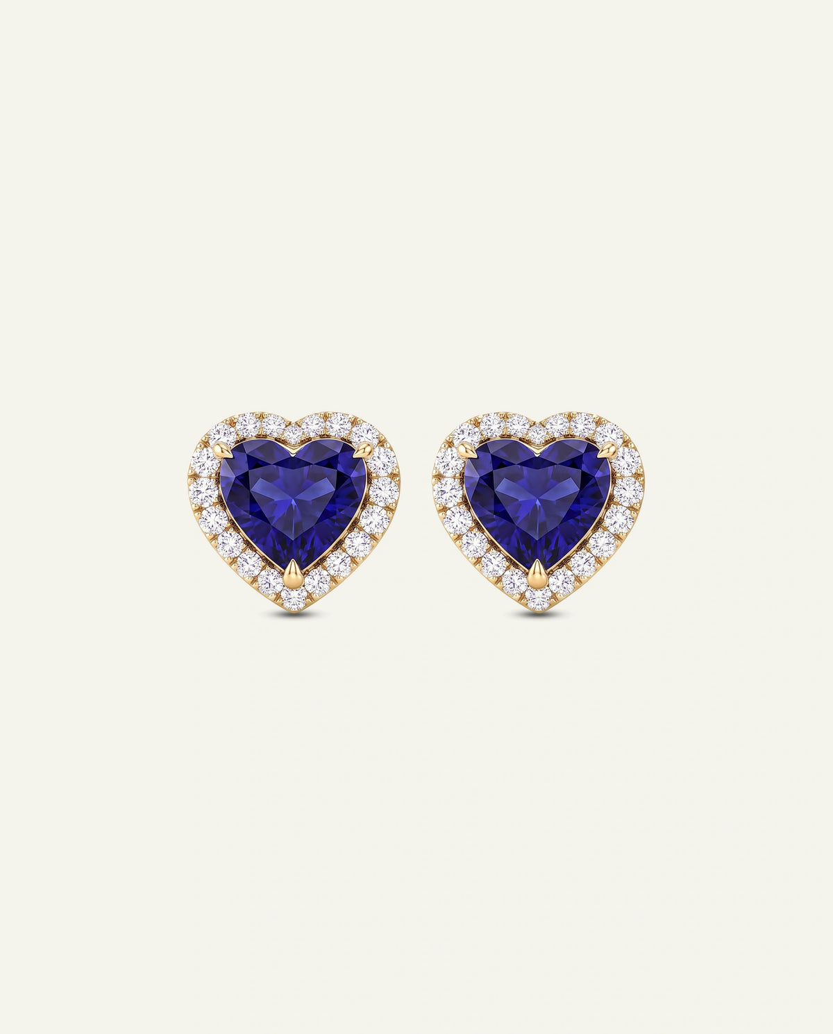 A pair of heart-shaped stud earrings featuring deep blue heart-shaped gemstones surrounded by a halo of small clear stones in a yellow gold setting, presented on a light cream background.