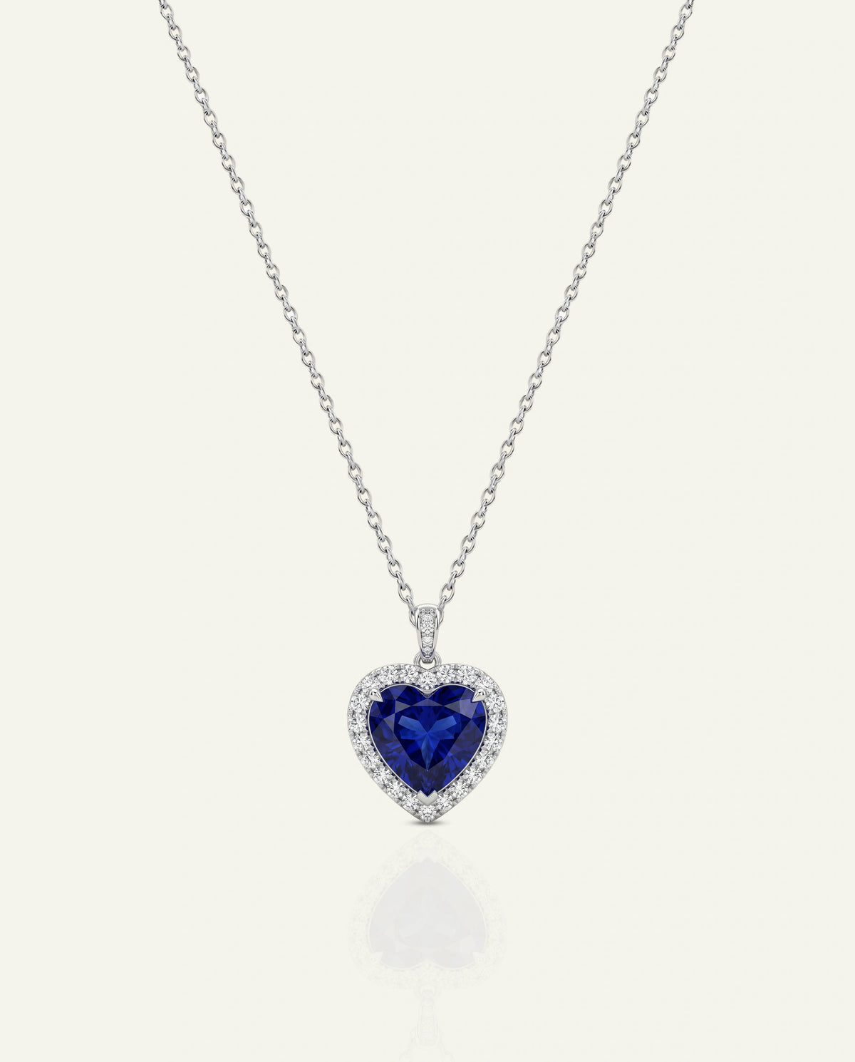 A heart-shaped pendant necklace featuring a deep blue heart-shaped gemstone surrounded by a halo of small clear stones, suspended on a delicate silver chain against a light cream background.