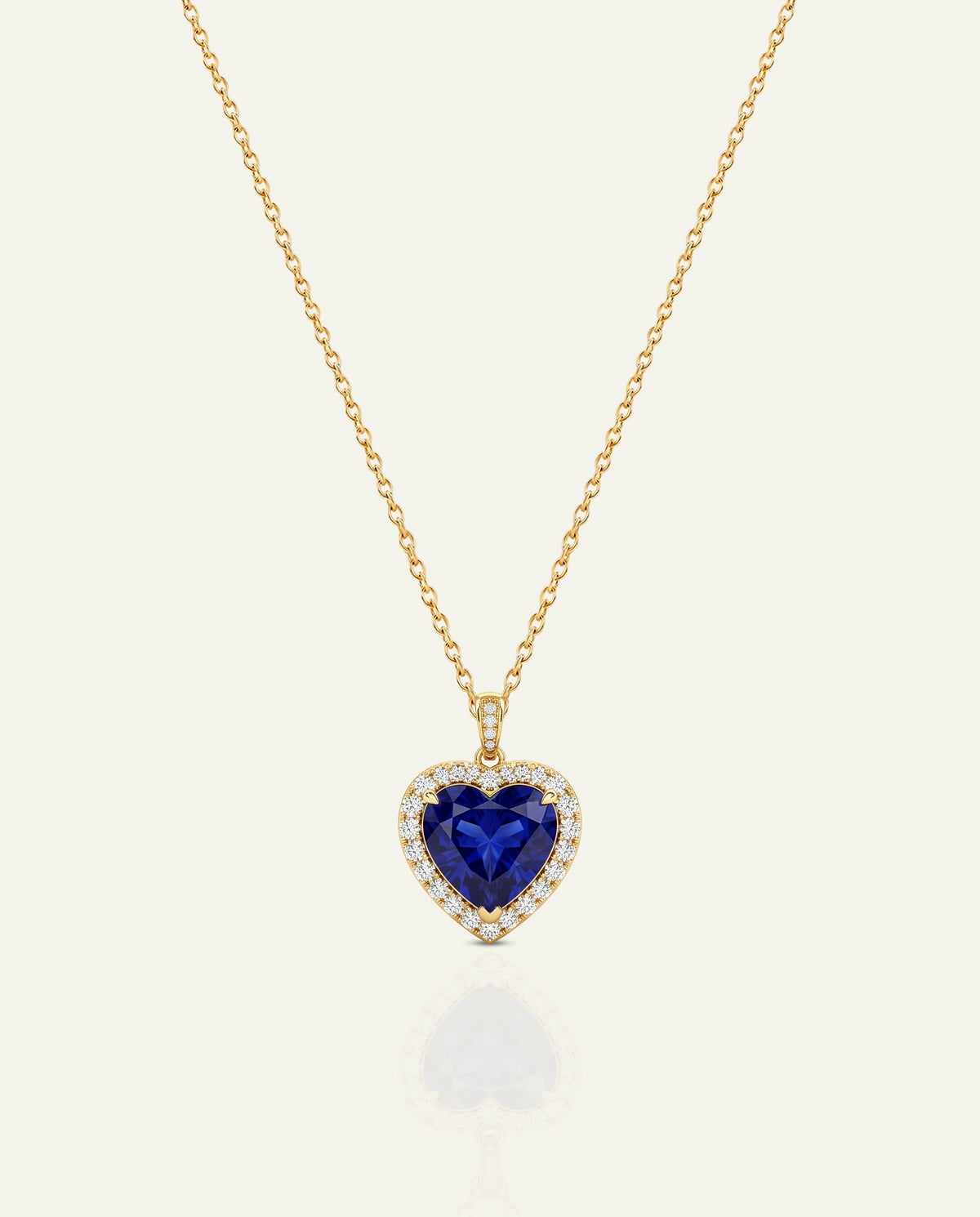 A gold heart-shaped pendant necklace featuring a deep blue heart-shaped gemstone surrounded by small clear stones, suspended on a delicate gold chain against a light cream background.