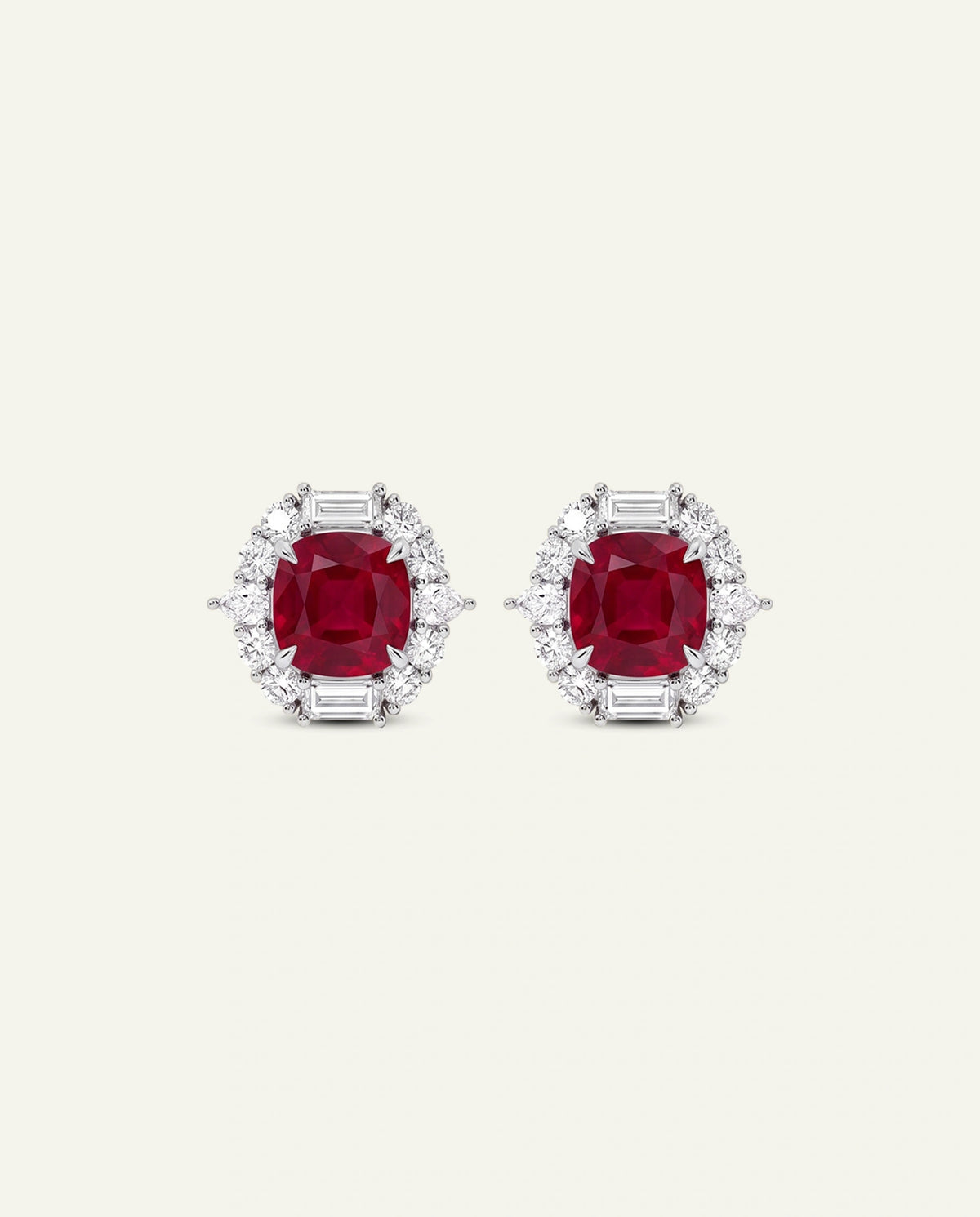 A pair of white gold stud earrings featuring vibrant cushion-cut red rubies framed by a halo of baguette and round clear diamonds, set against a light cream background.