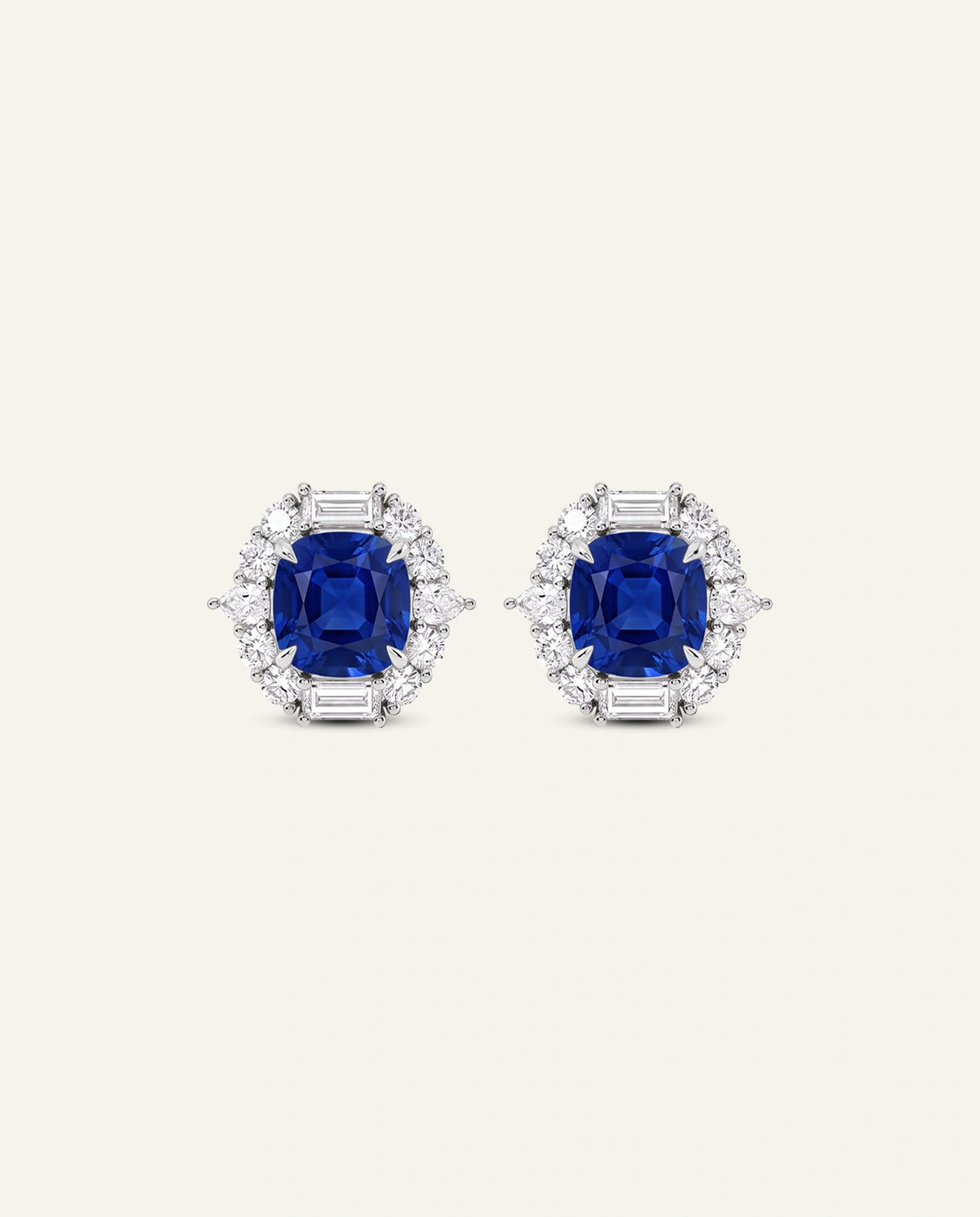 A pair of oval stud earrings featuring a central cushion-cut deep blue sapphire surrounded by a halo of round and baguette-cut clear stones, set in a silver-toned metal on a light cream background.
