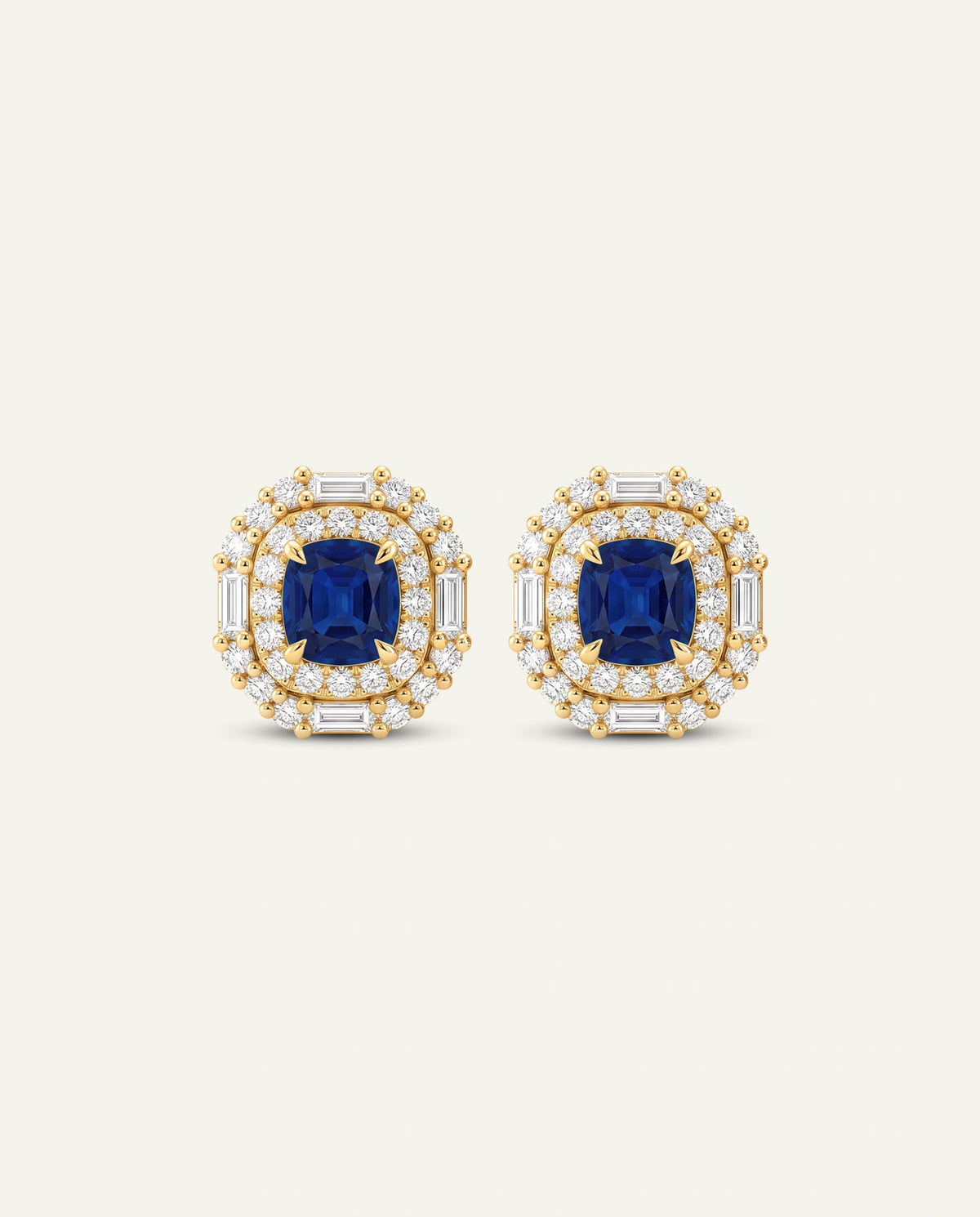 A pair of yellow gold stud earrings featuring a central cushion-cut blue sapphire surrounded by a double halo of small round diamonds and baguette diamonds, set against a light cream background.