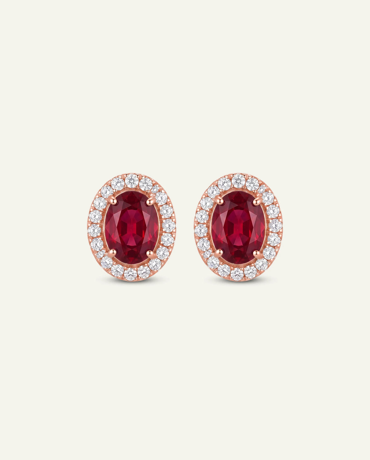 A pair of oval stud earrings featuring a central deep red ruby stone surrounded by a halo of small clear stones in a rose gold setting, shown on a light cream background.