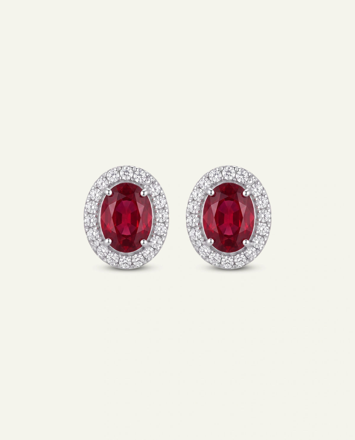 A pair of oval stud earrings featuring a central deep red ruby stone surrounded by a halo of small clear stones in a silver-toned setting, shown on a plain light beige background.