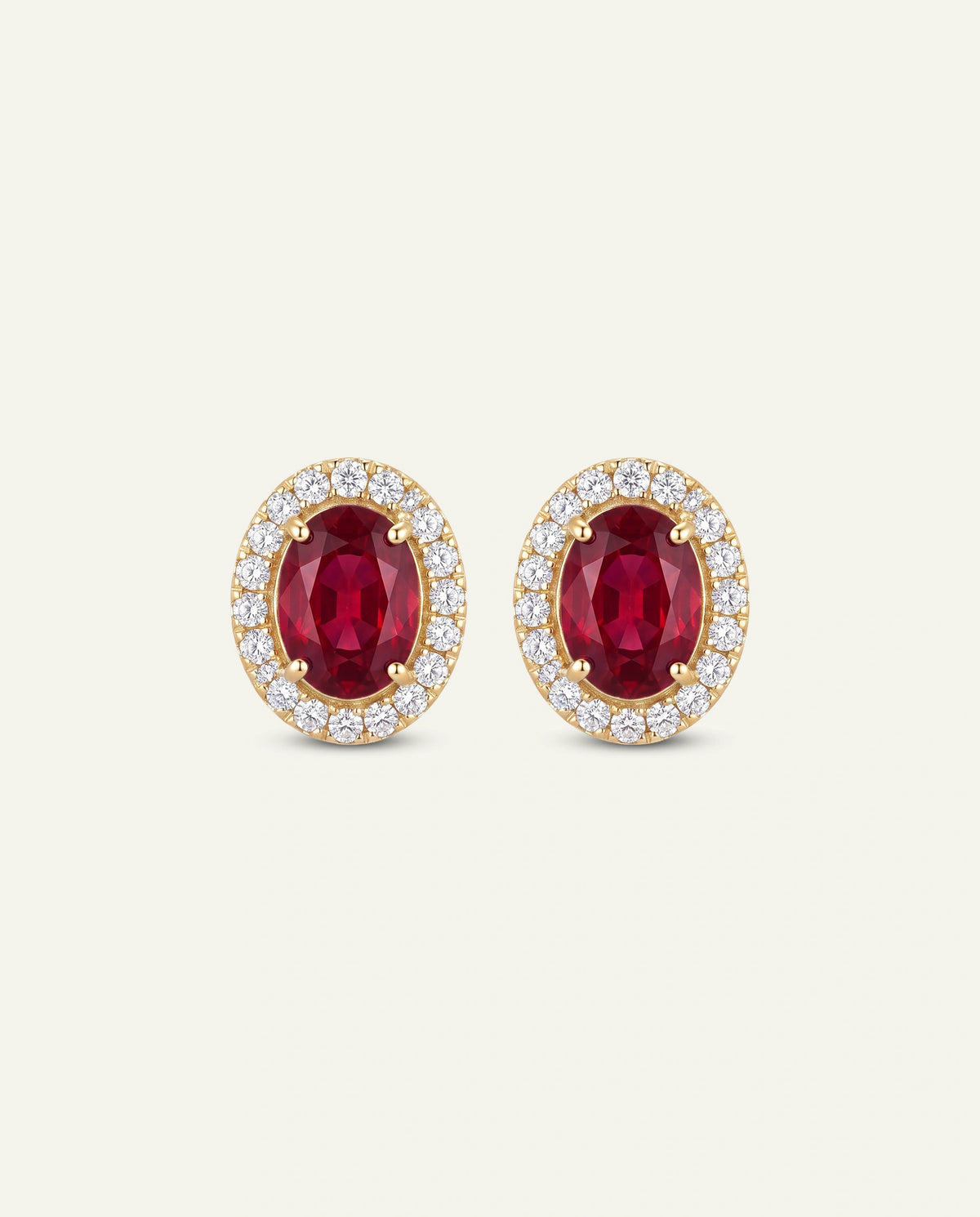 A pair of oval yellow gold stud earrings featuring a central oval-cut deep red ruby center stone surrounded by a halo of small round clear stones, set against a light cream background.