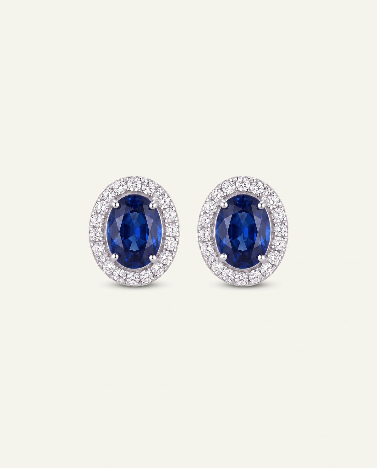 A pair of oval stud earrings featuring deep blue sapphires surrounded by a halo of small clear stones in a silver-toned setting, presented on a light cream background.