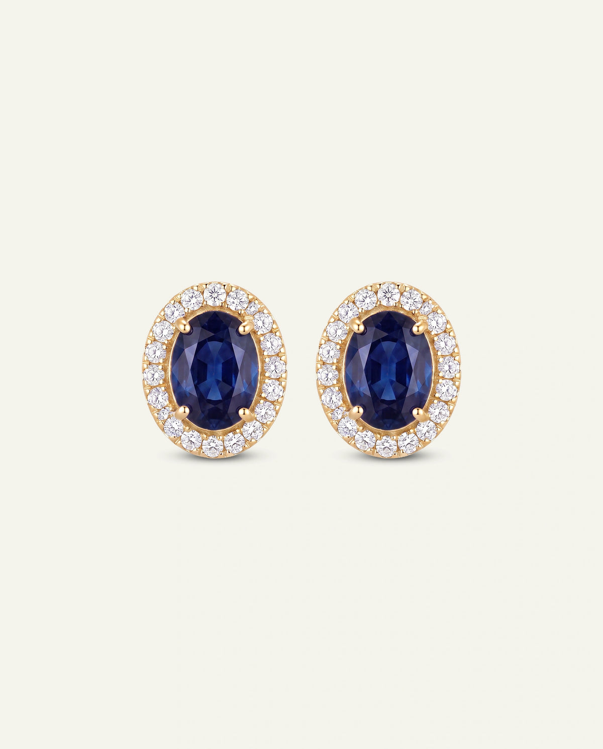 A pair of oval stud earrings featuring deep blue sapphires surrounded by a halo of small clear stones in a yellow gold setting, presented on a light cream background.