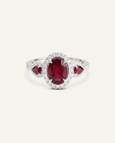 A silver ring featuring a large oval-cut deep red ruby center stone surrounded by a halo of small clear stones, with two smaller heart-shaped red ruby stones on the shoulders, set against a light cream background. - alternate view