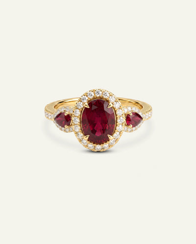 A yellow gold ring featuring a large oval-cut deep red ruby center stone, surrounded by a halo of small clear pavé stones, with two smaller marquise-cut red ruby side stones on the shoulders, set against a light off-white background.