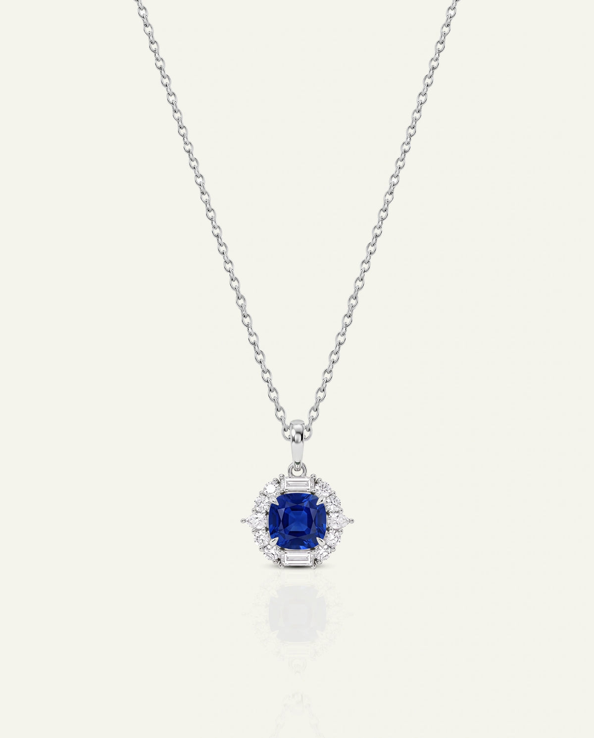 A white gold chain necklace featuring a cushion-cut deep blue sapphire pendant framed by a halo of round brilliant diamonds and baguette accent diamonds, shown on an off-white background.