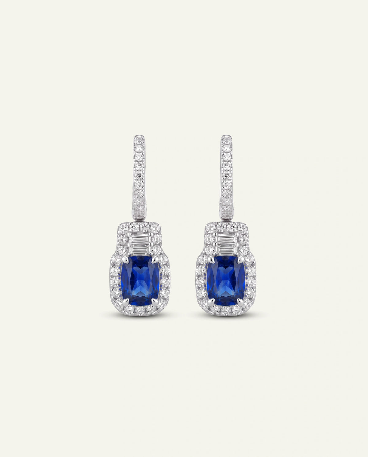 A pair of silver drop earrings featuring a rectangular emerald-cut blue sapphire center stone surrounded by a halo of small clear stones, with a vertical pavé-set silver post above.
