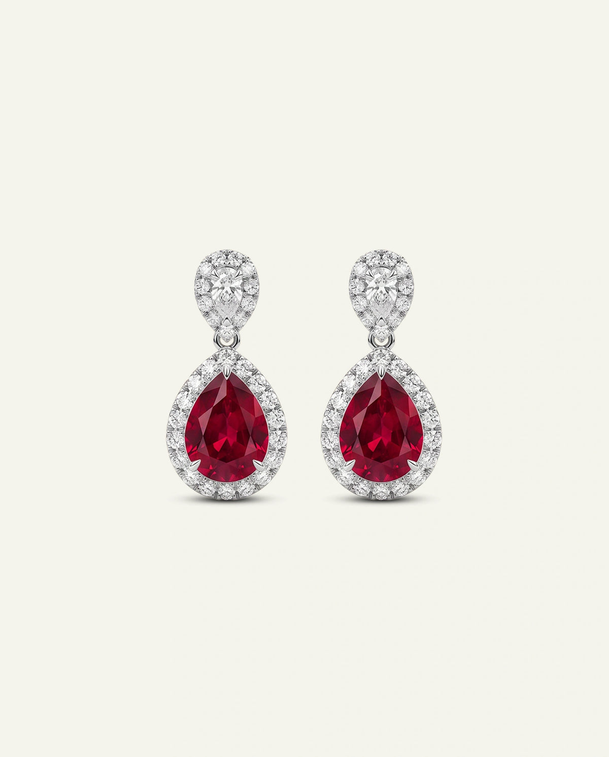 A pair of teardrop-shaped drop earrings featuring large round-cut deep red gemstones surrounded by small clear stones in a silver-toned setting, shown on a light cream background.