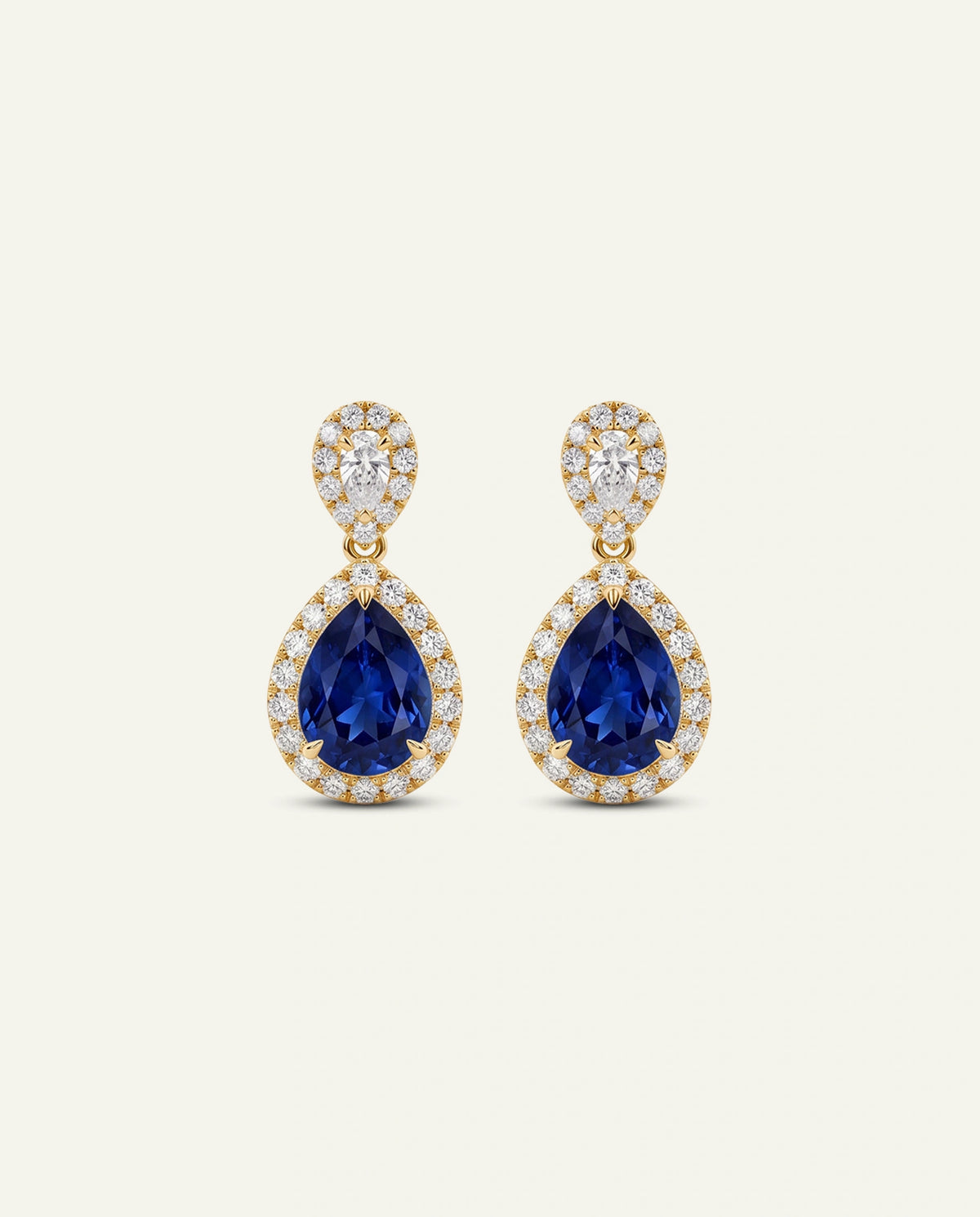 A pair of gold drop earrings featuring pear-shaped deep blue sapphires surrounded by small clear stones, with a smaller round cluster of clear stones above, set against a light cream background.