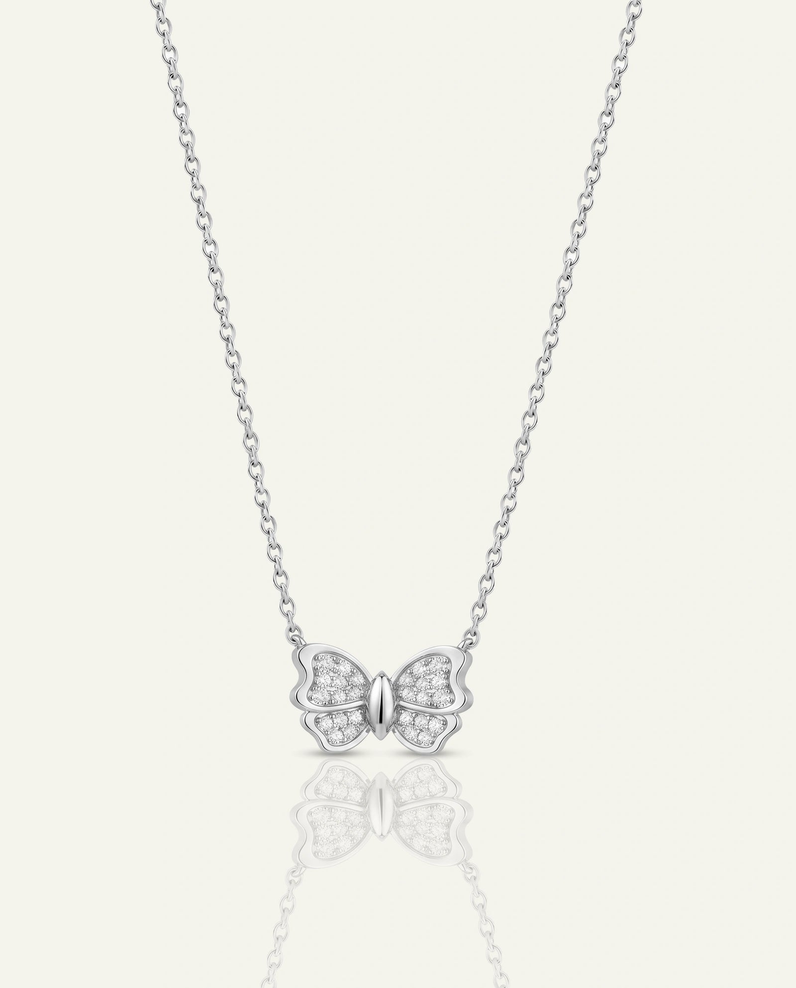A delicate silver chain necklace featuring a butterfly pendant paved with small clear stones, centered on a light cream background with a soft reflection below.